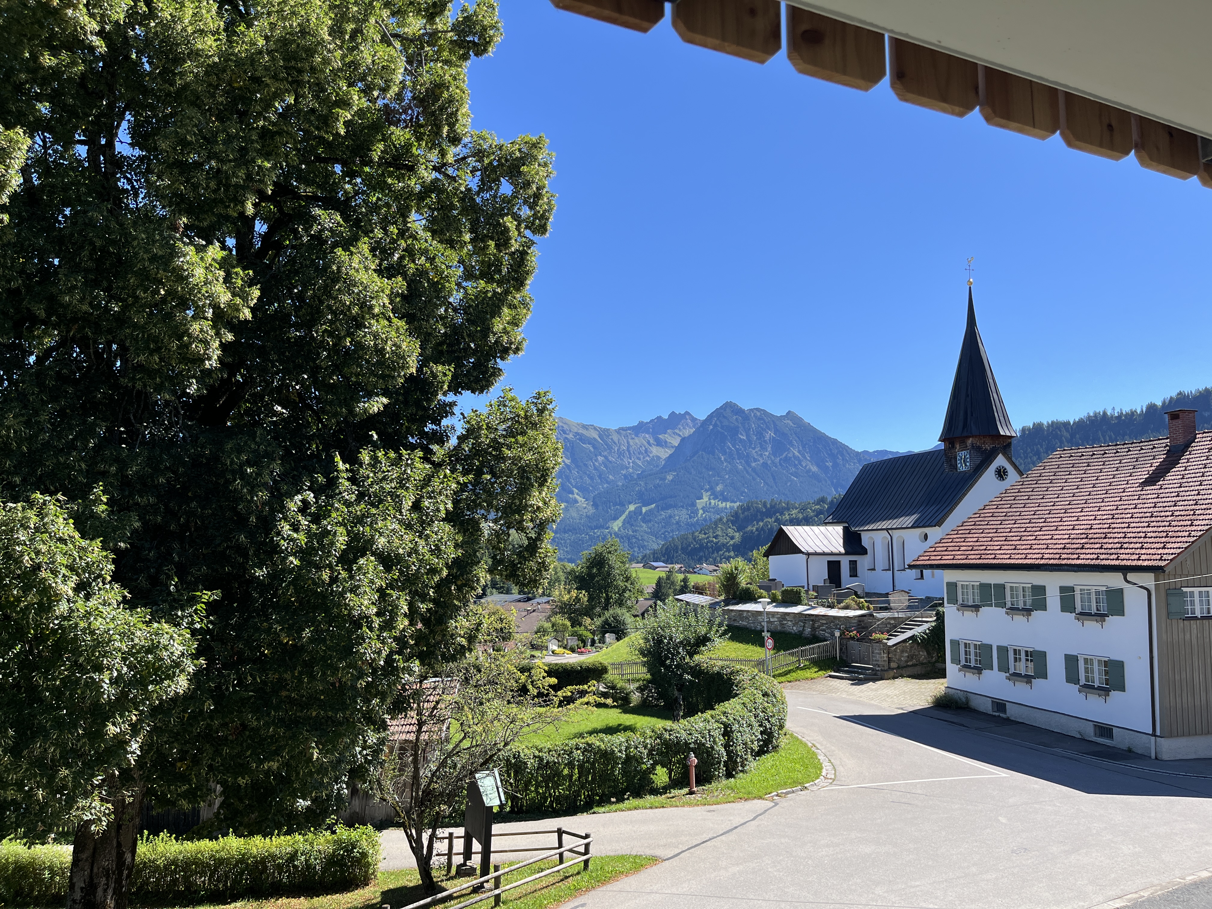 Eine malerische Landschaft mit einer Kirche, einem charmanten Dorf und Bergen im Hintergrund. Der Himmel ist klar und blau, umgeben von grüner Natur.