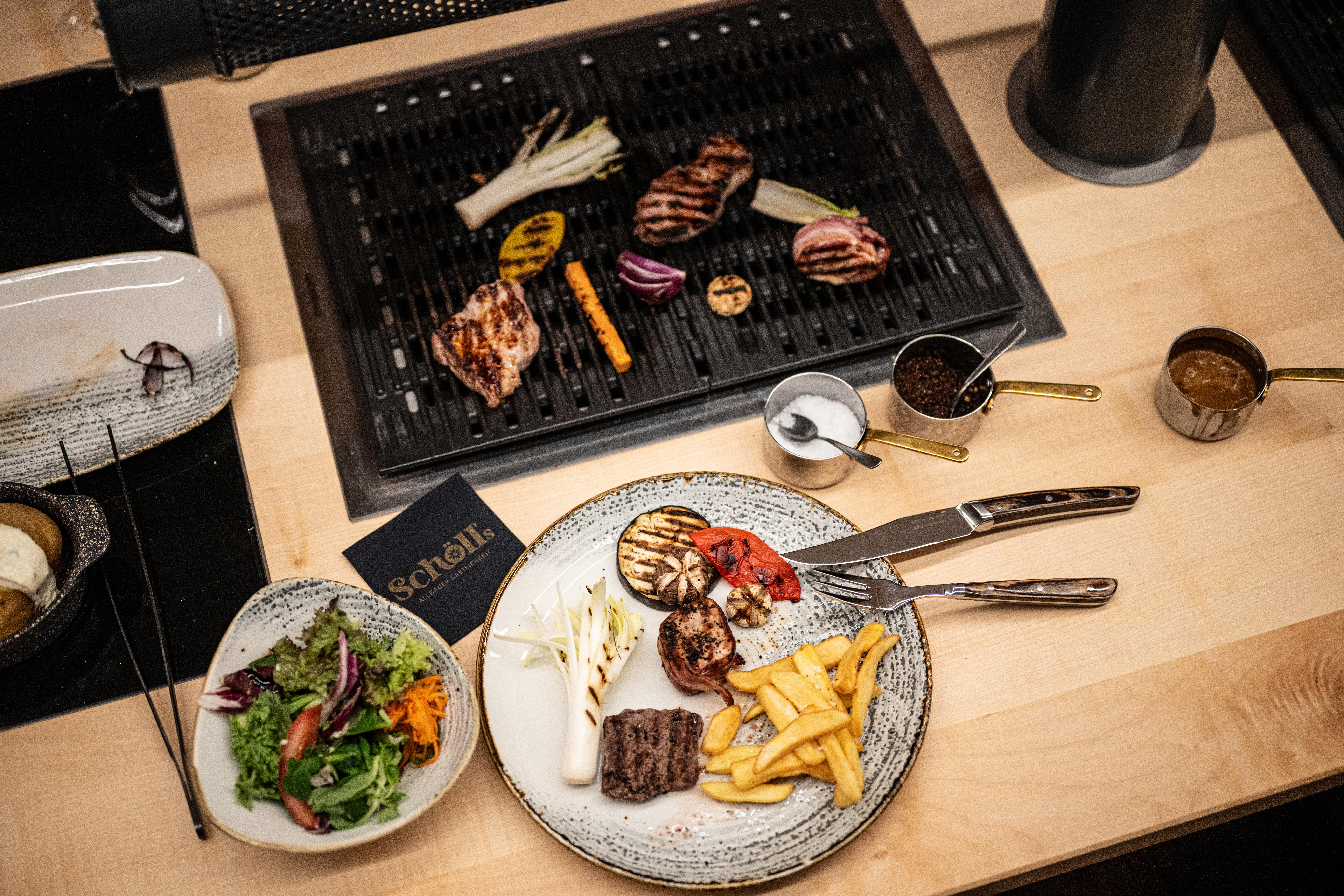 Tischgrill in Holzplatte eingelassen, Fleisch, Gemüse, Pommes Frites, Ofenkartoffel und verschiedenen Soßen