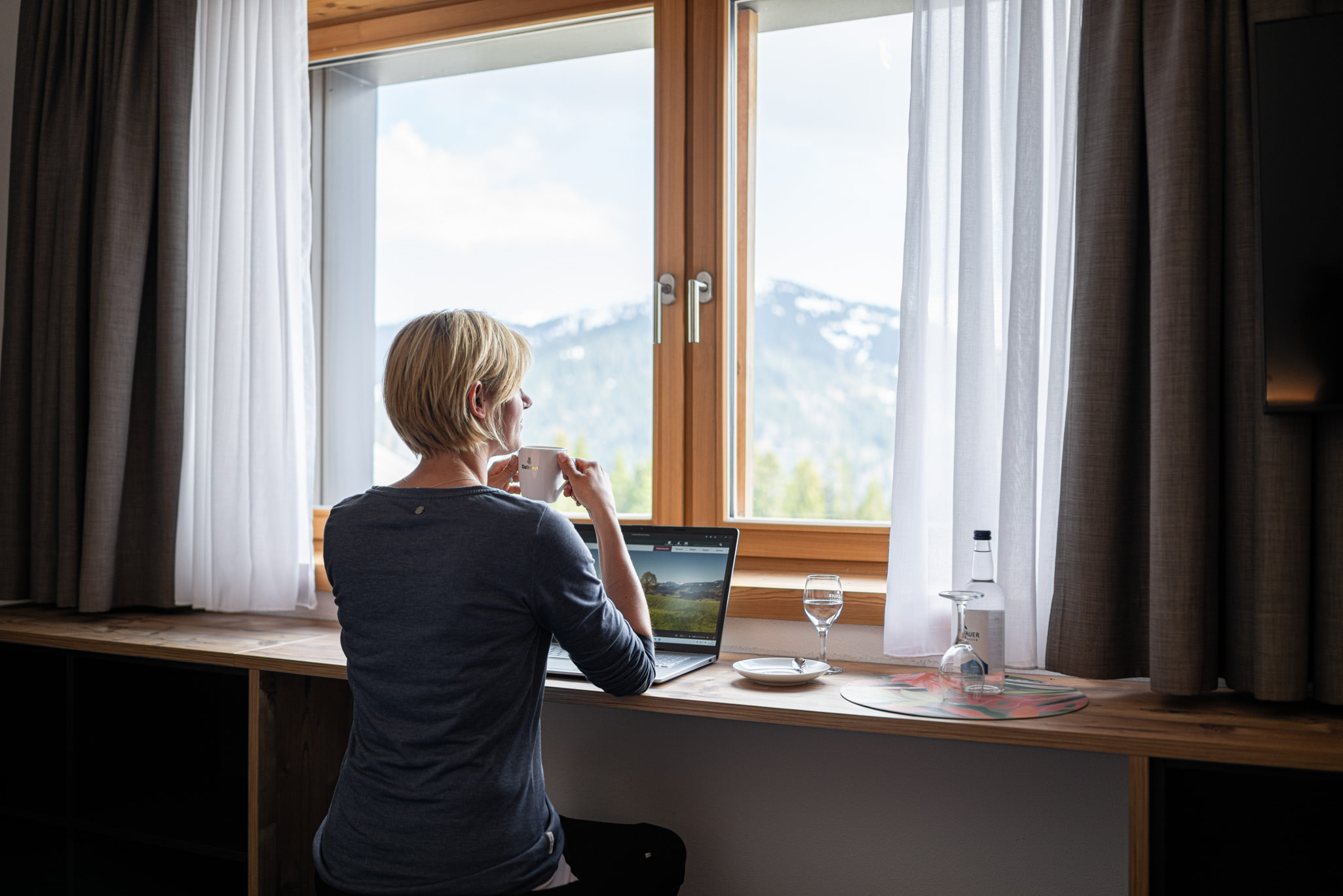 Frau am Fenster mit Laptop, Kaffee, Wasserflasche. Blick auf die Allgäuer Berge in Balderschwang, blauer Himmel.