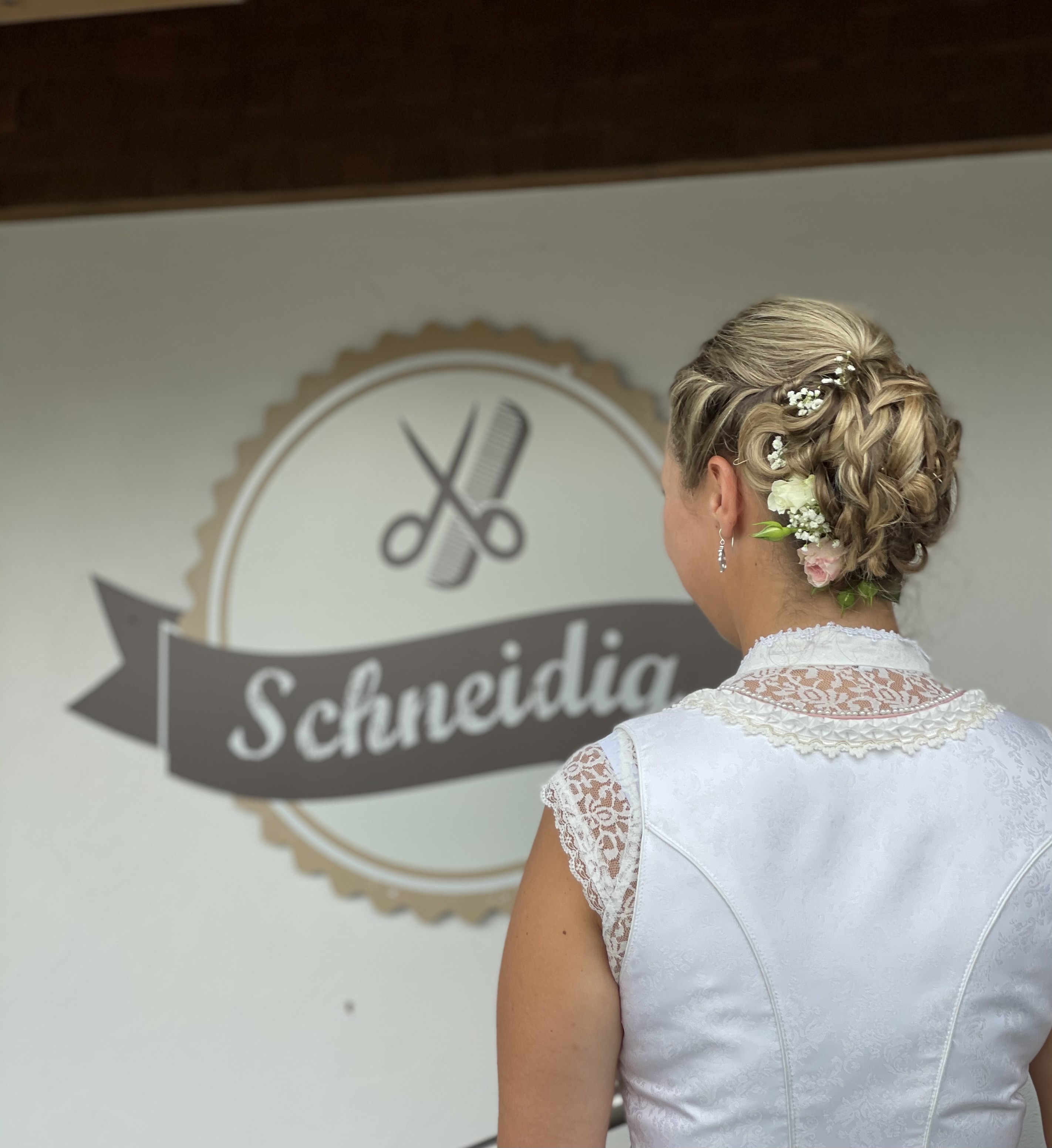Brautfrisur von Schneidig Elegante Braut in Rückenansicht, das kunstvolle Brautkleid und die perfekte Hochzeitsfrisur im Vordergrund. Im Hintergrund das Logo von Friseur Schneidig.