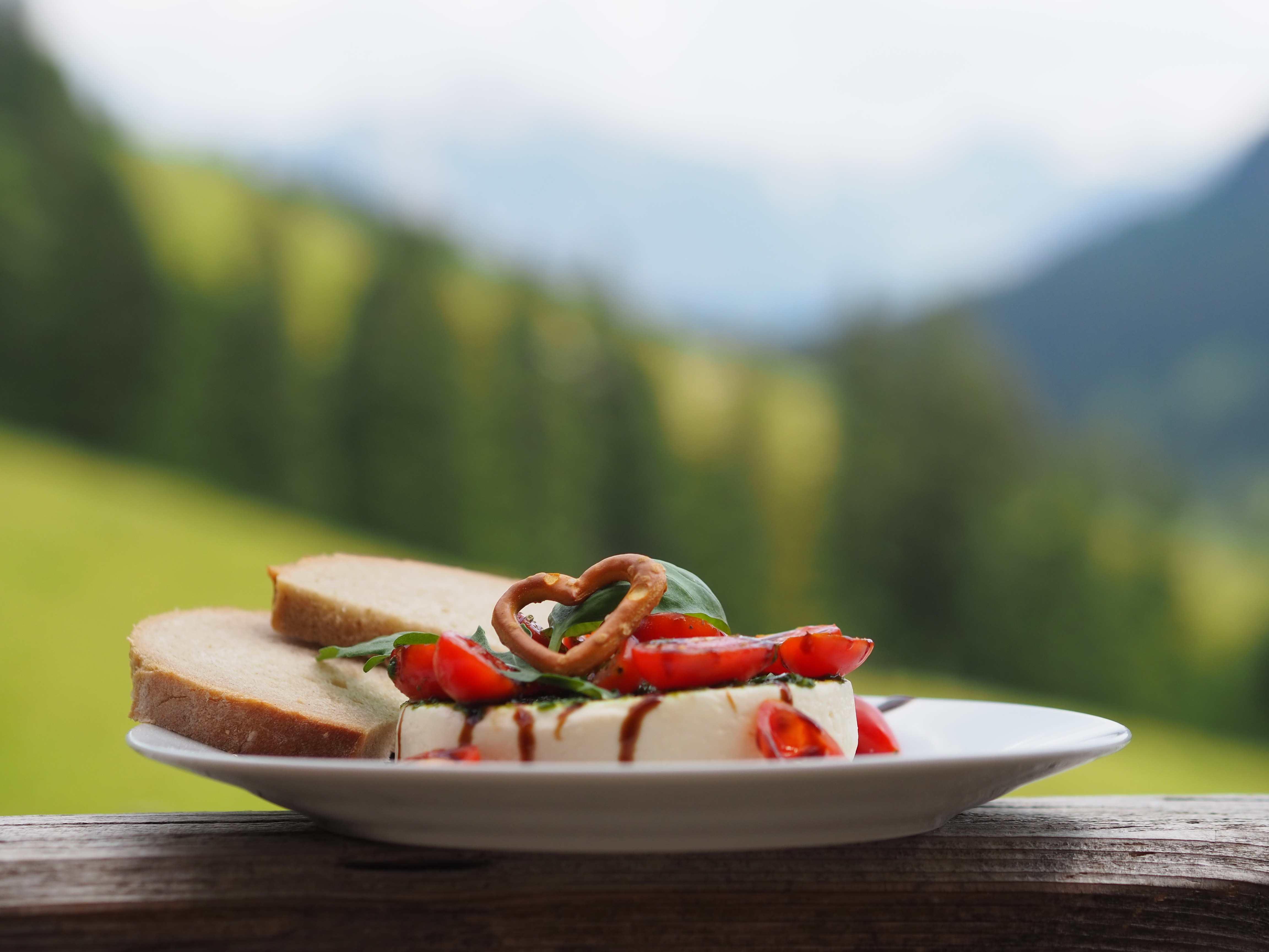 Auf Teller angerichteter Weichkäse mit Tomaten, Pesto und Brotscheiben auf Veranda. Im Hintergrund Alplandschaft. Ein Rezept einer Alpe in den Hörnerdörfern im Allgäu.
