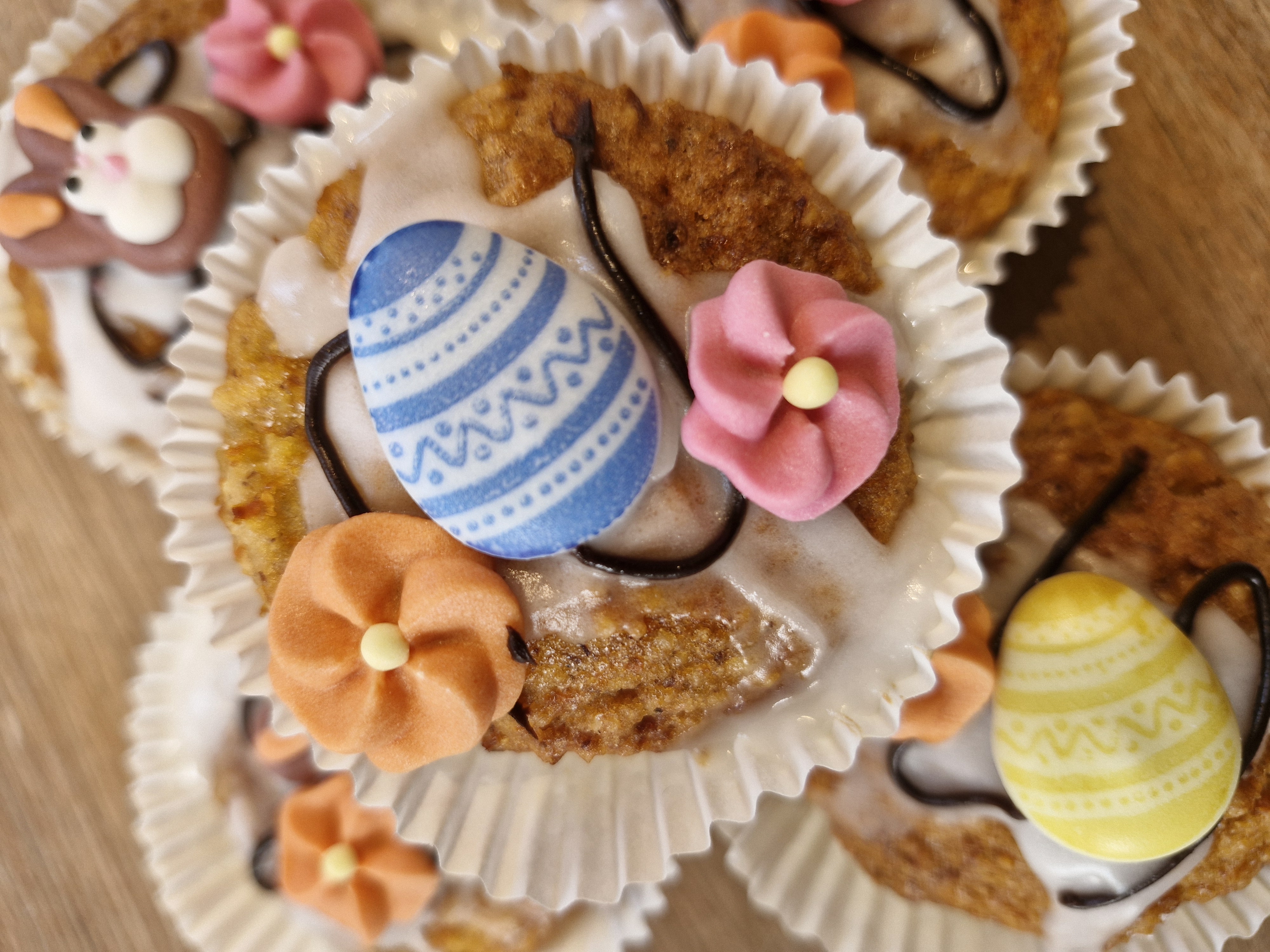 Hausgemachte Oster-Muffins mit Karottenteig, Zuckerguss, bunten Zucker-Eiern und Frühlingsblüten dekorativ auf Holz angerichtet.