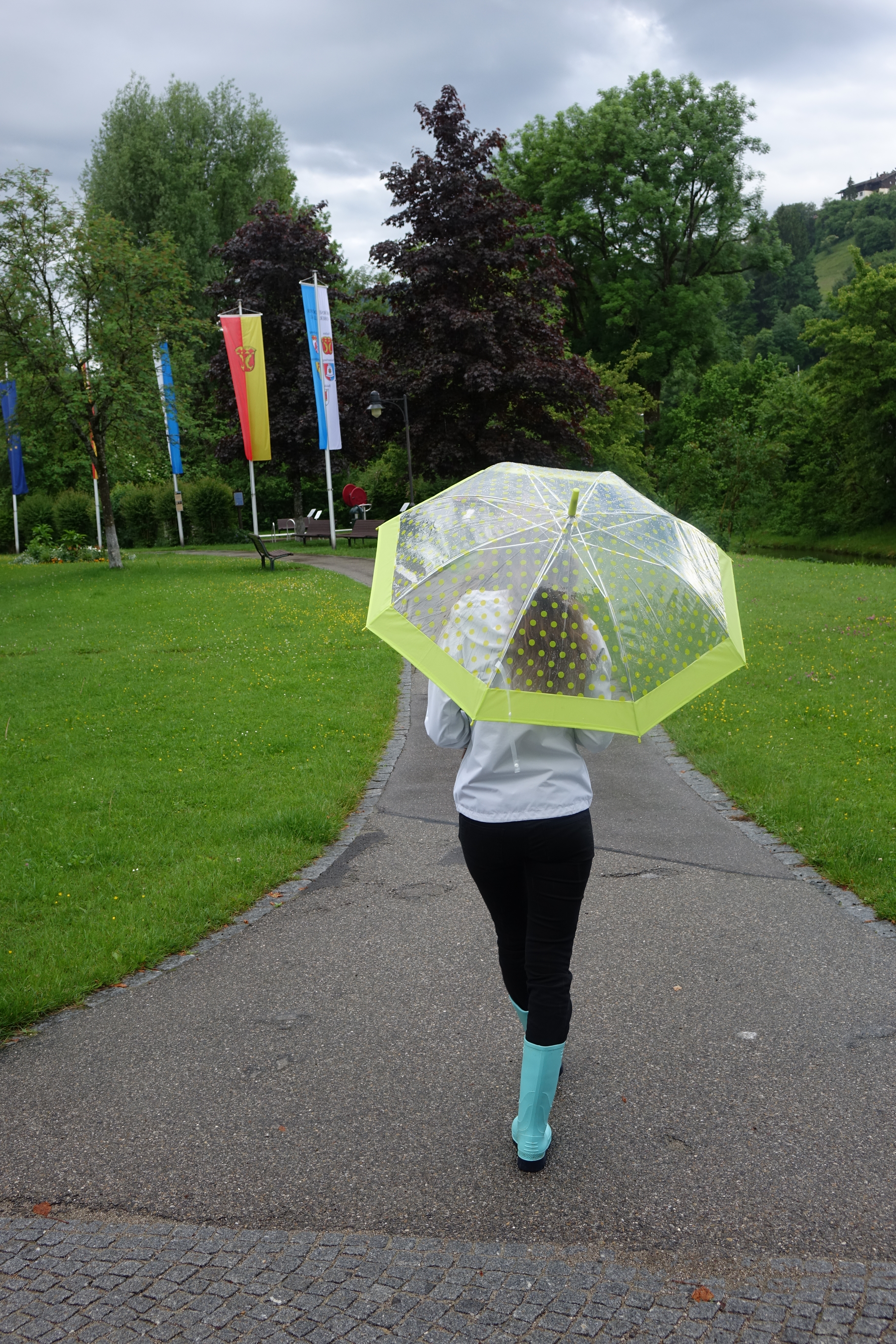 Frau mit Regenschirm und Gummistiefeln spaziert im herrlich grünen Kurpark in Fischen im Allgäu