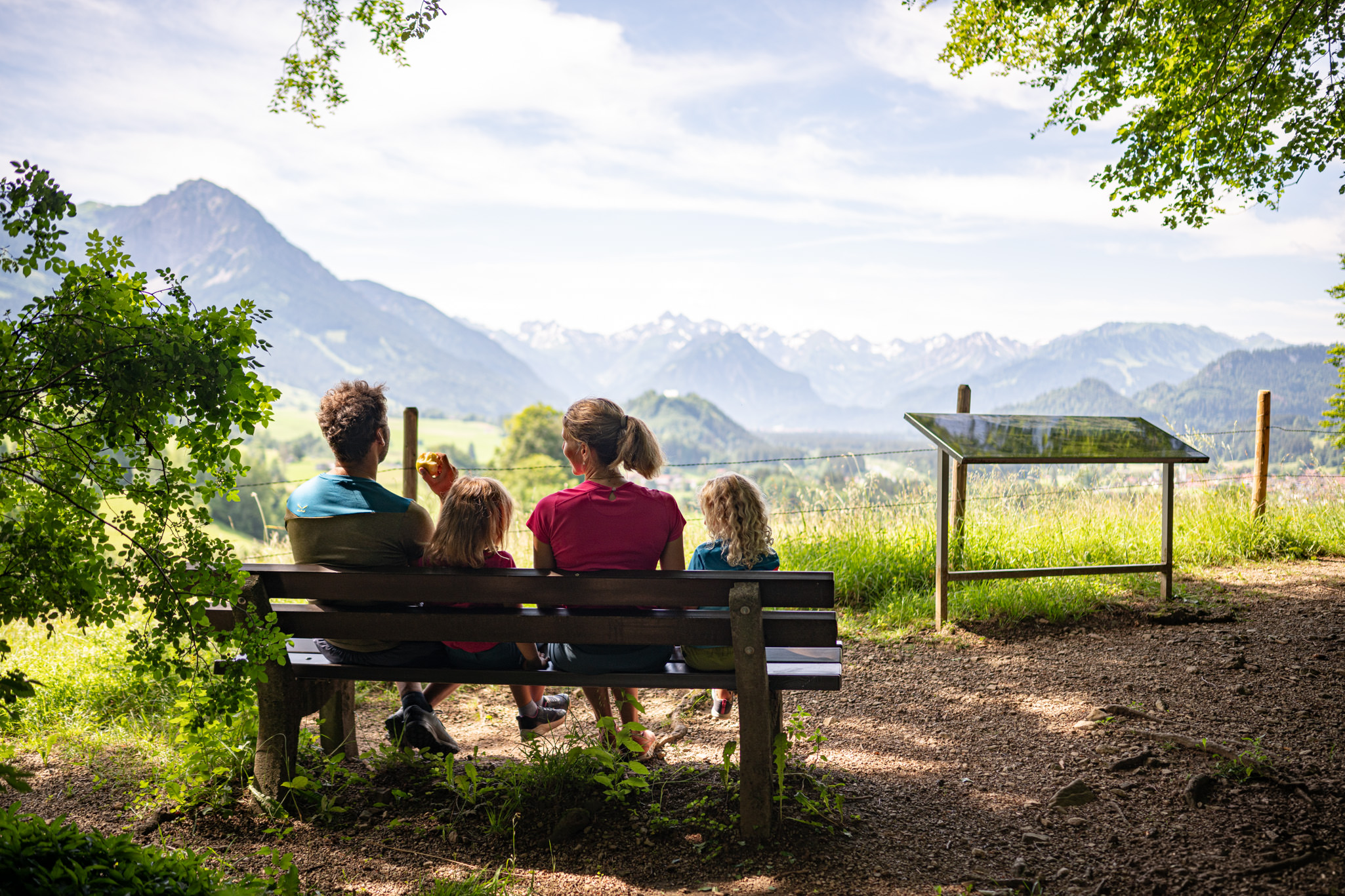 Eltern mit 2 Kindern sitzen auf Aussichtsbank mit Blick auf das Bergpanorama, umgeben von Bäumen.