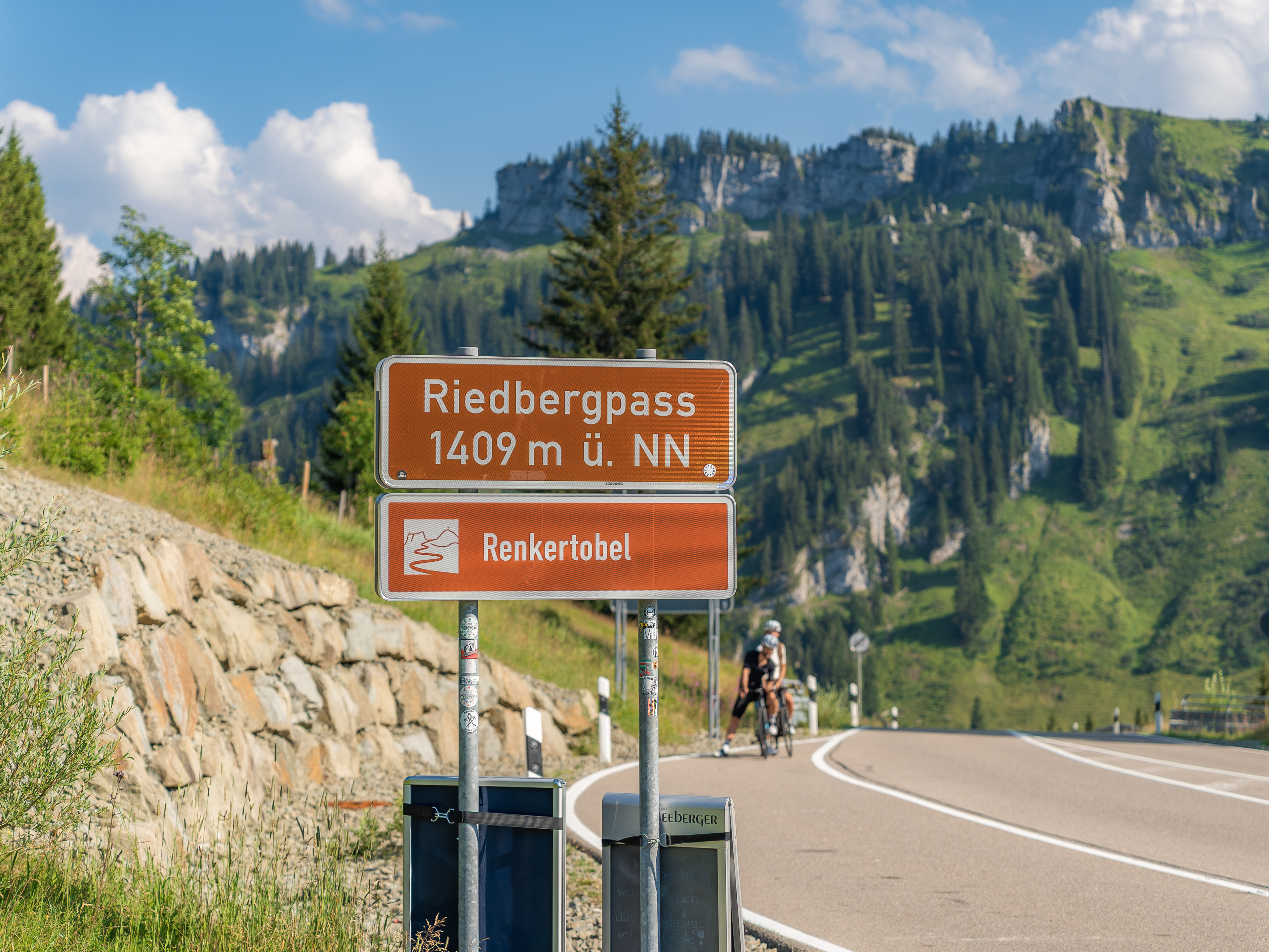 Riedbergpass 1409m ü. NN und Renkertobel Straßenschilder mit Berglandschaft und Radfahrern im Hintergrund.