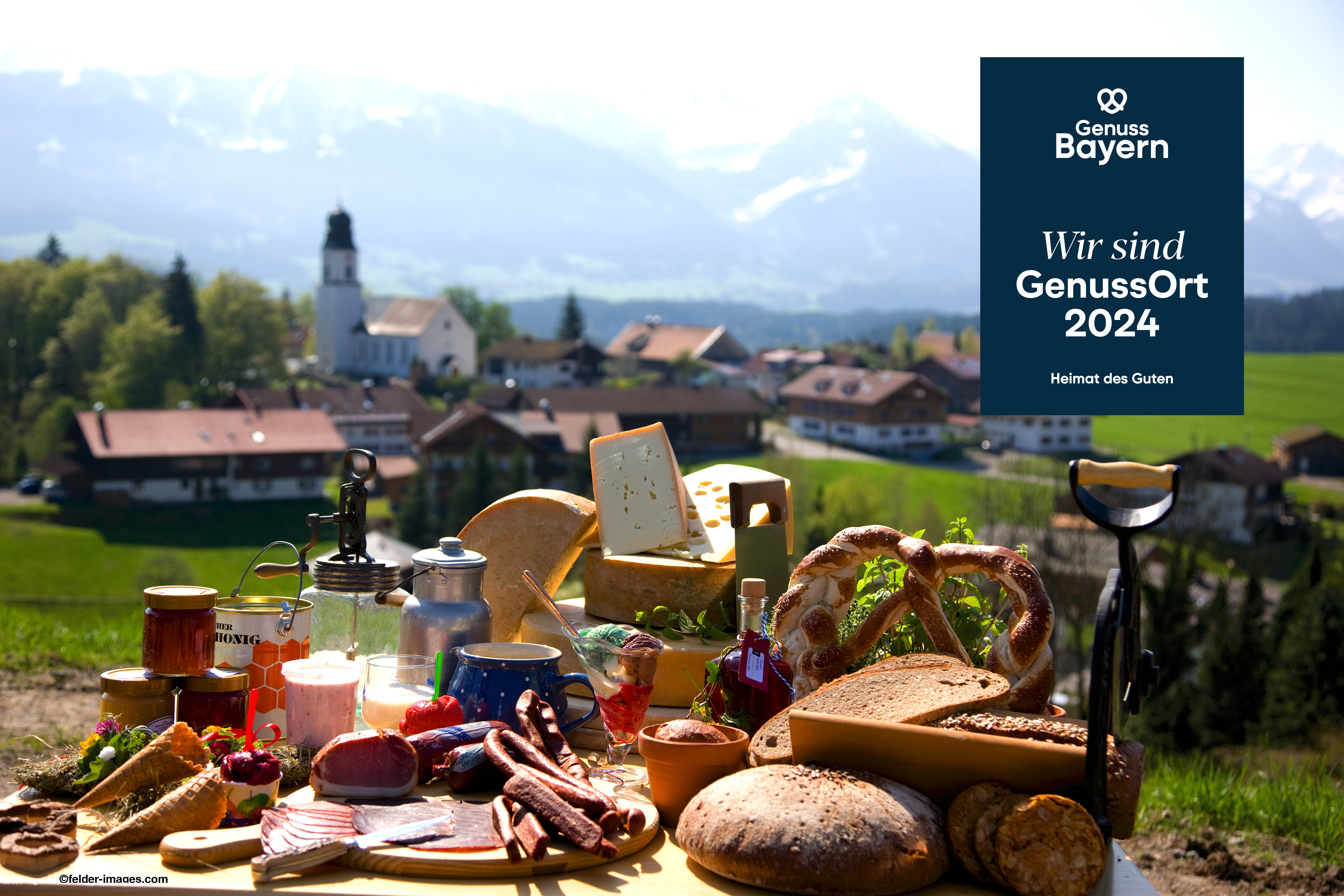 Ofterschwang "Gutes vom Dorf" - GenussOrt2024 Ein Tisch mit regionalen Spezialitäten aus Ofterschwang vor Dorf und Bergpanorama. Rechts oben das Logo "Genuss Bayern. GenussOrt 2024. Heimat des Guten".