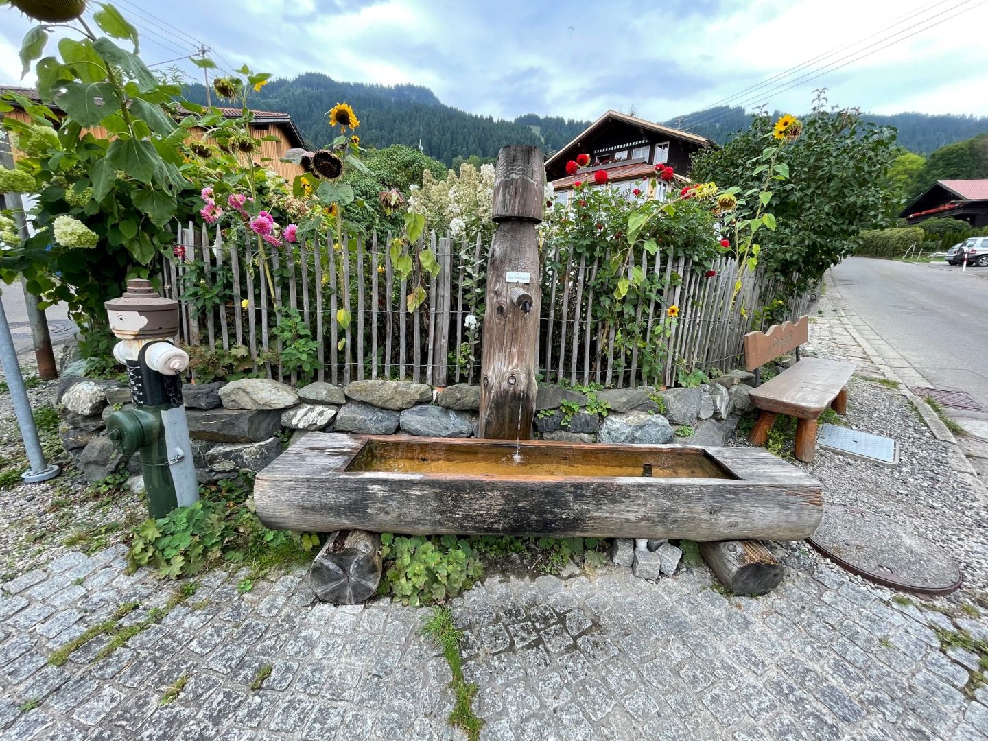 Holzbrunnen vor Bauernzaun mit Bauerngarten, an der alten Schule in Bolsterlang.