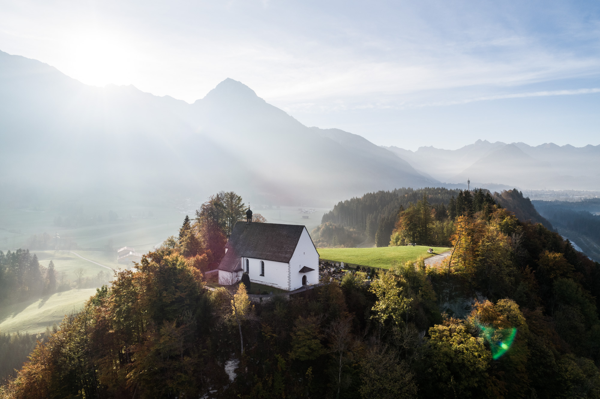 Im Sonnenlicht präsentiert sich die Schöllanger Burgkirche, umrahmt von Bäumen und den Gipfeln der Berge.