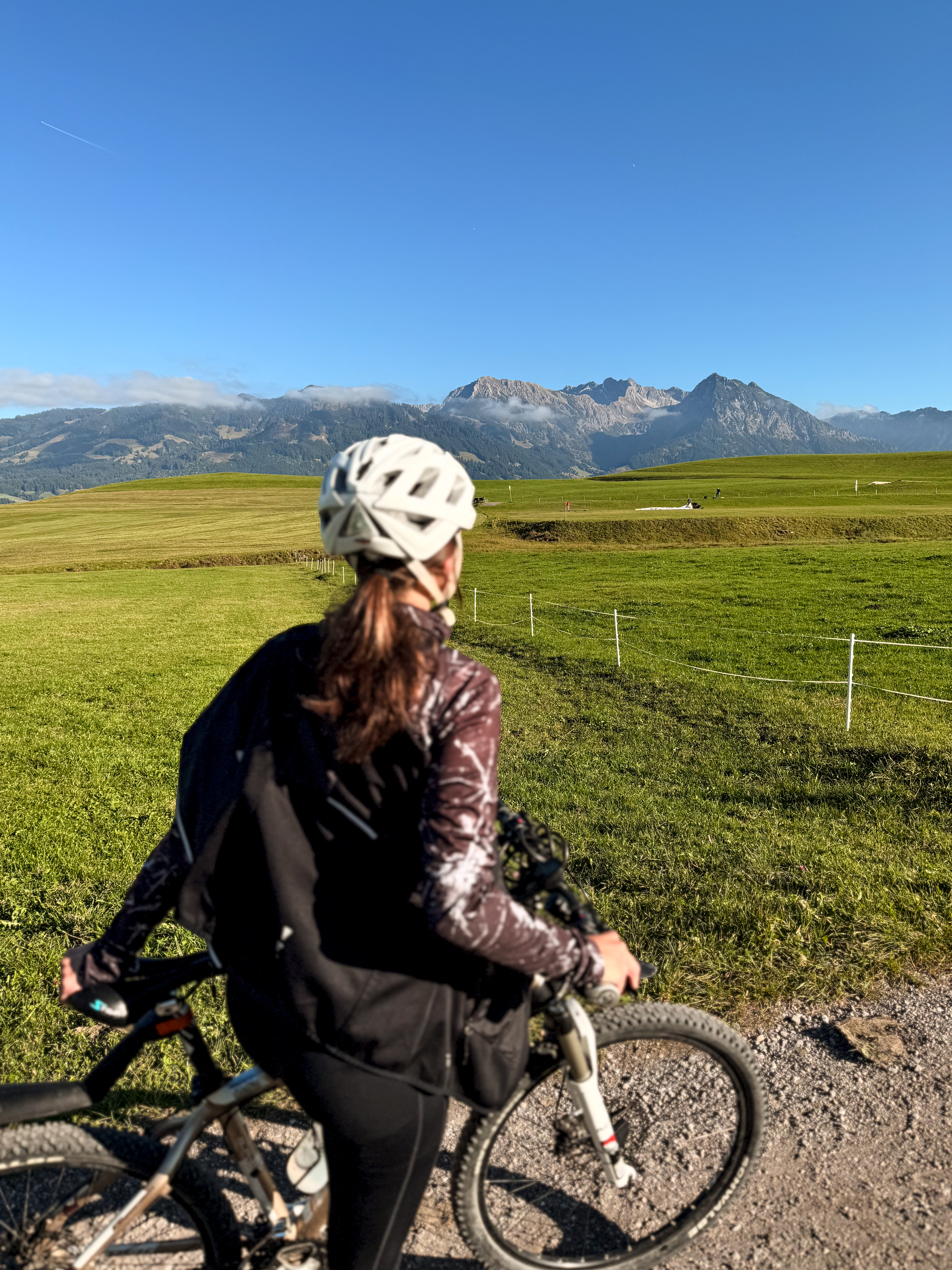 Eine Radfahrerin blickt von hinten auf eine weite grüne Wiese und eine Bergkette unter blauem Himmel.