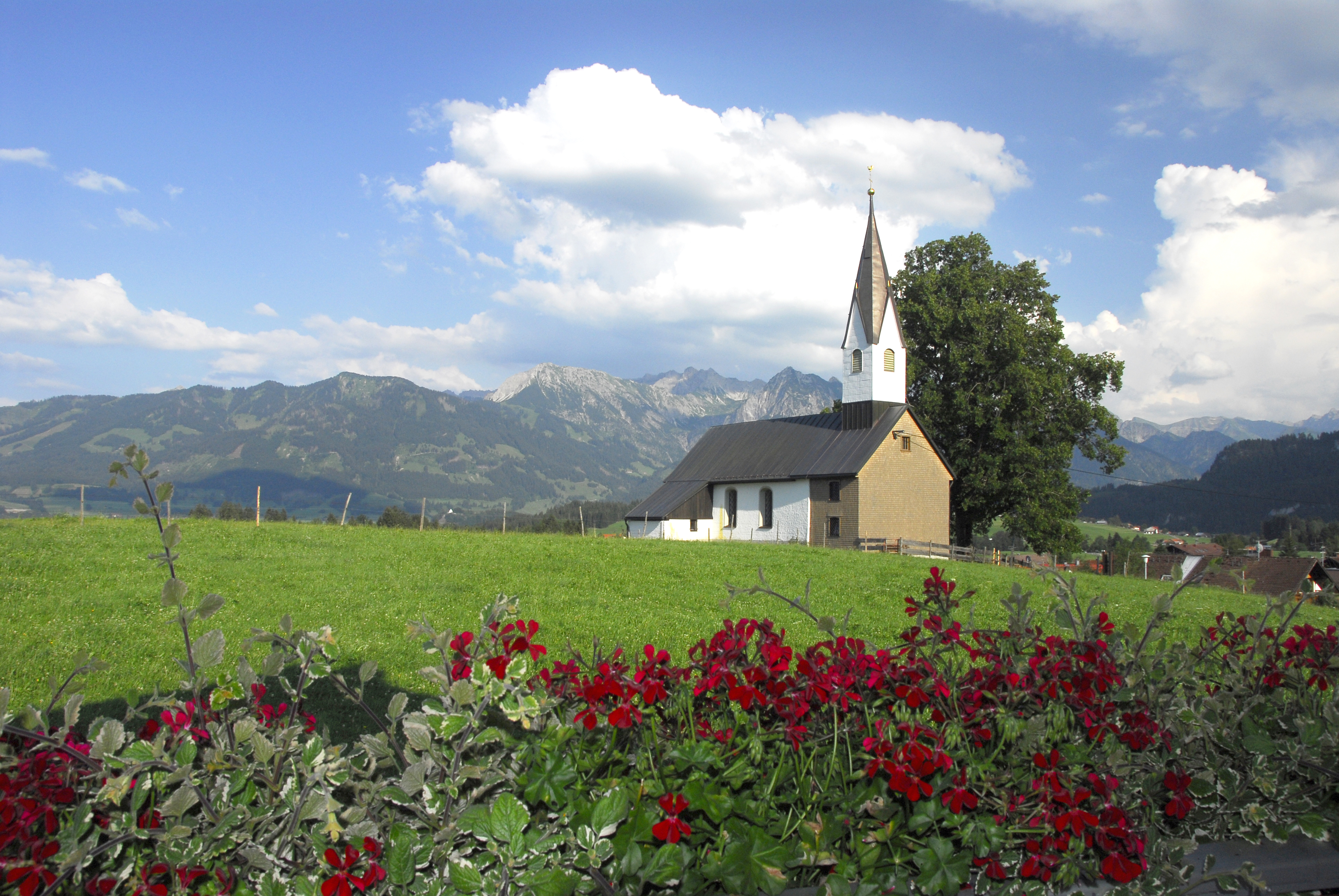 Kapelle in Bolsterlang in den Hörnerdörfern im Allgäu. Im Hintergrund Berge, im Vordergrund Wiese und Blumen.
