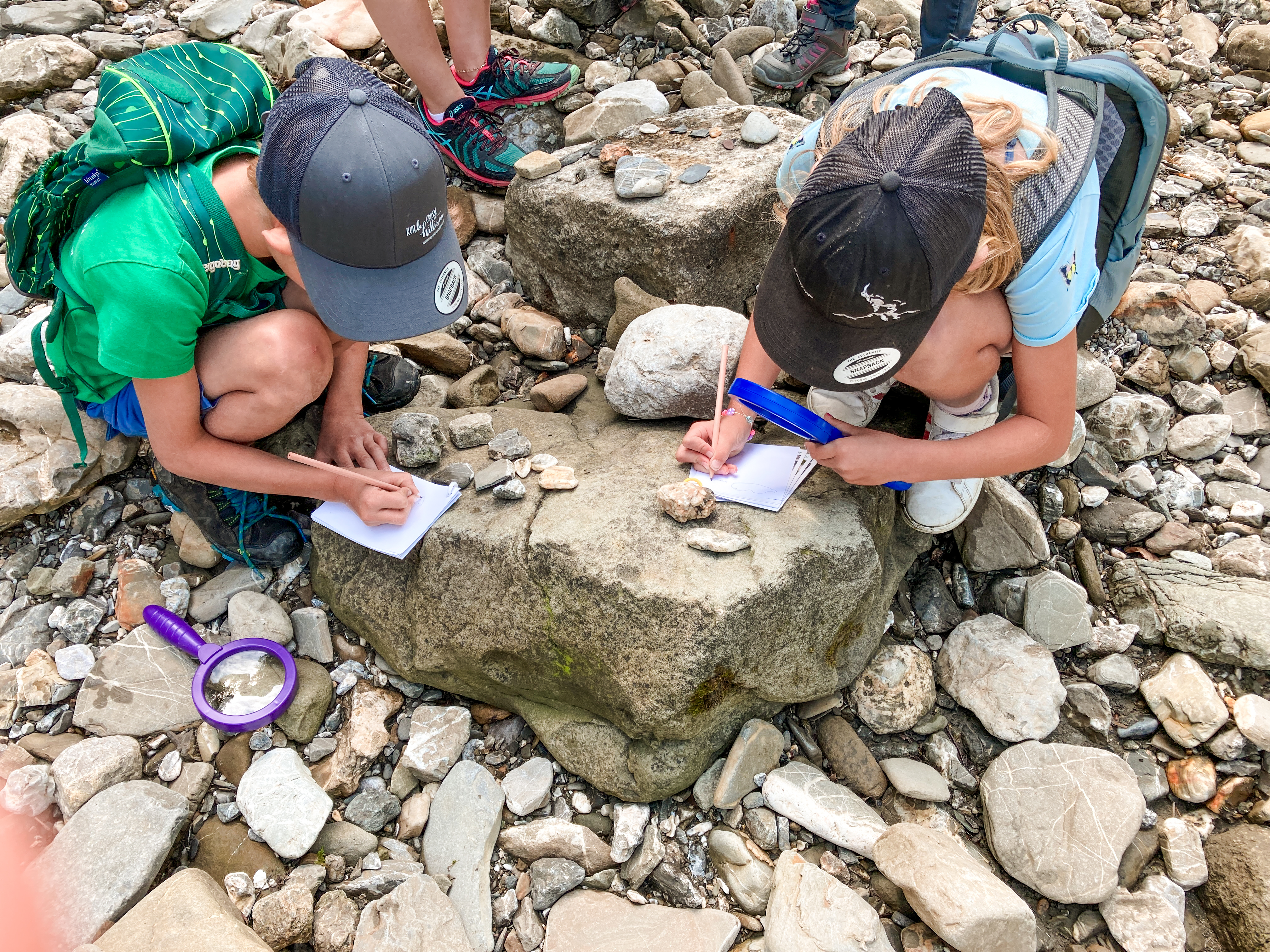  Zwei Kinder sitzen am Flussufer der Bolgenach und untersuchen mit Lupen Steine. Eines macht Notizen auf einem Papier.