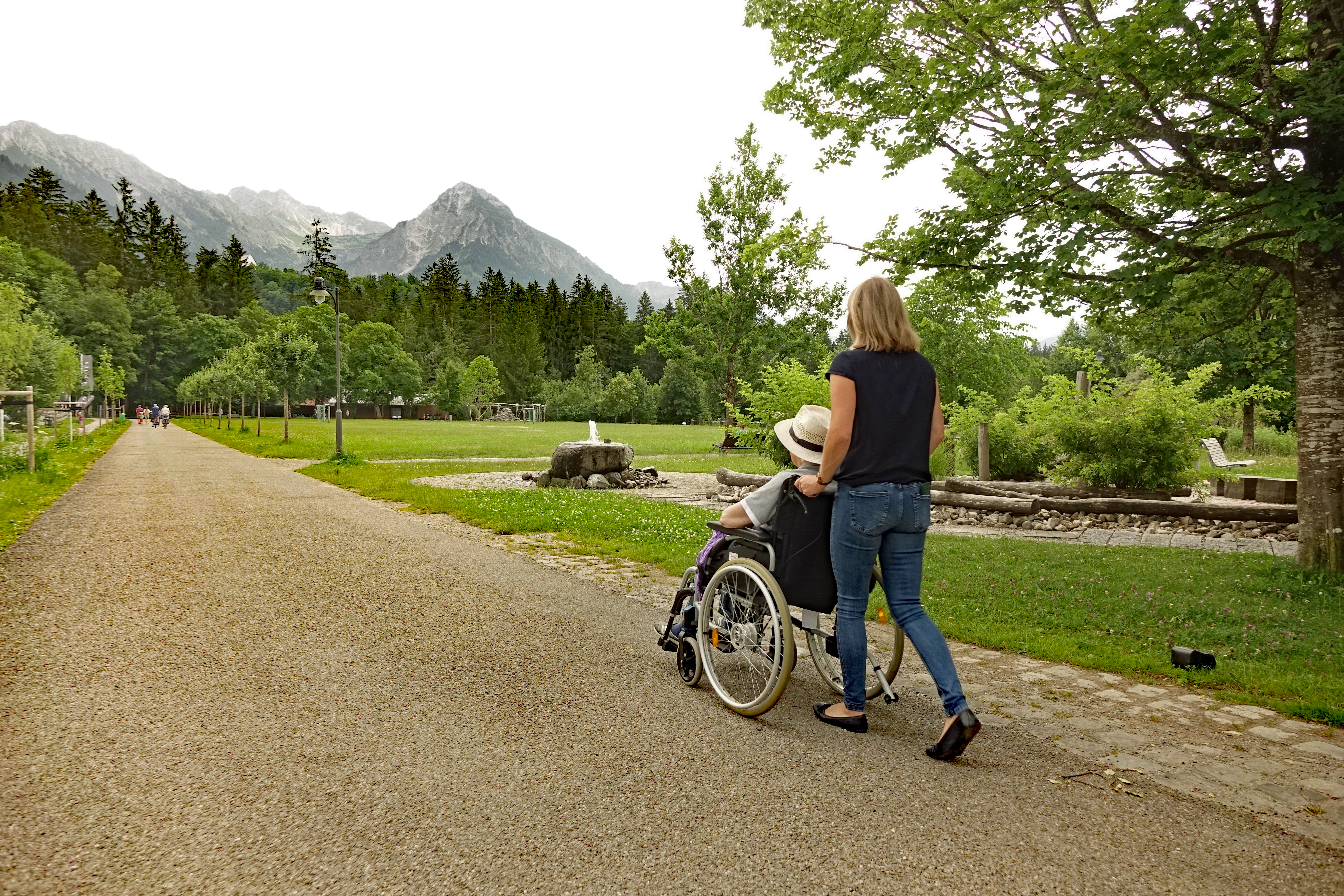 Frau schiebt Rollstuhl durch den Kurpark Fischen in den Hörnerdörfern im Allgäu. Hinten Berge, rechts Barfußpfad. 