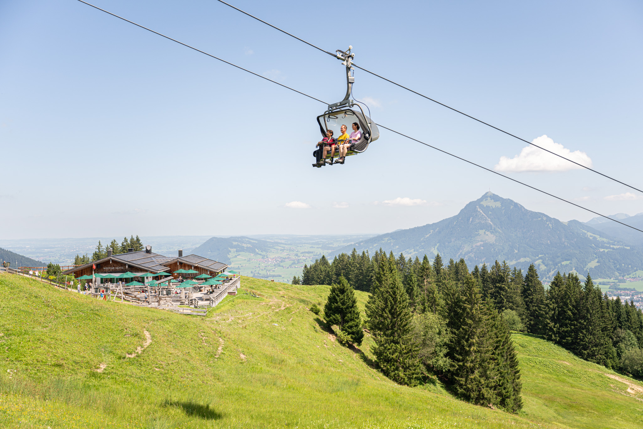 Sessellift schwebt über einer grünen Bergwiese mit Hütte und Bäumen vor blauem Himmel.