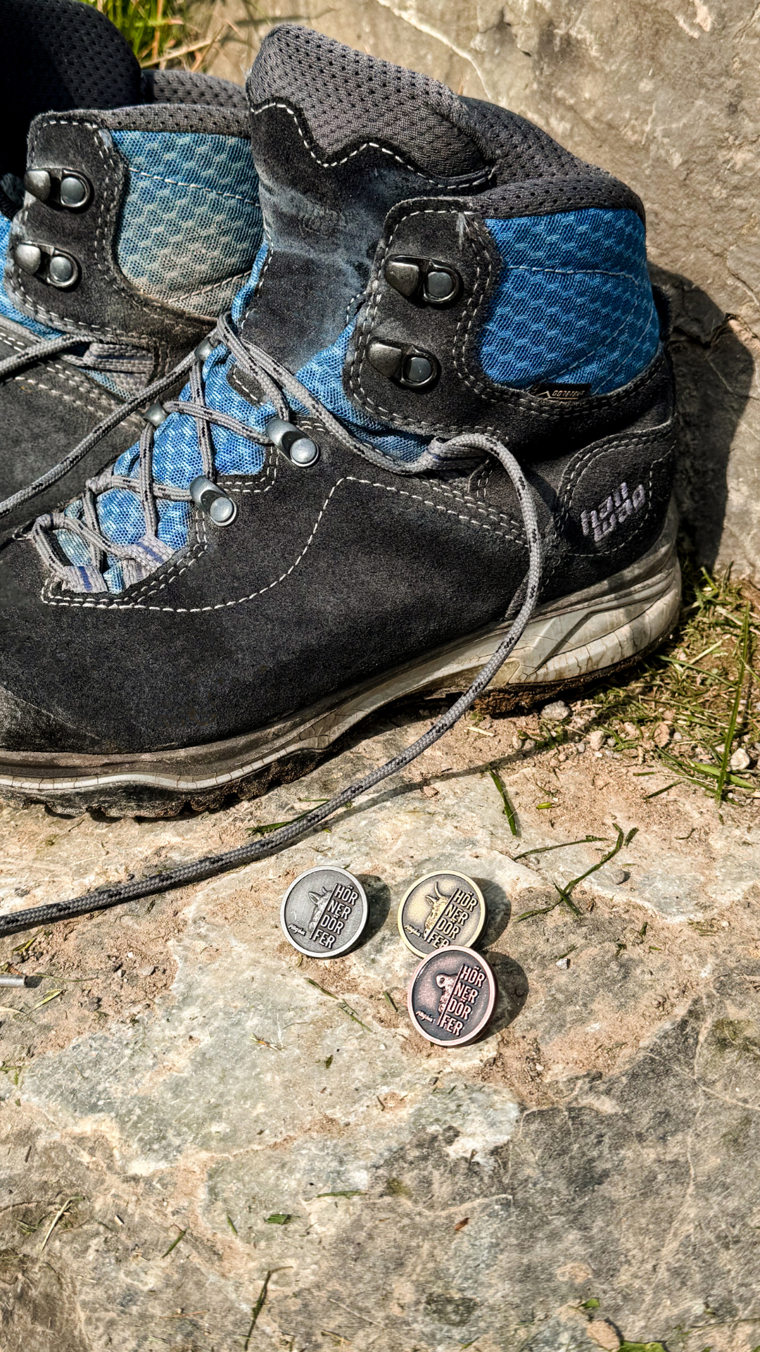 Ein Wanderschuh steht auf Felsen, davor liegen drei runde Metallanstecker mit Tier-Prägung und der Aufschrift "Hörnerdörfer".
