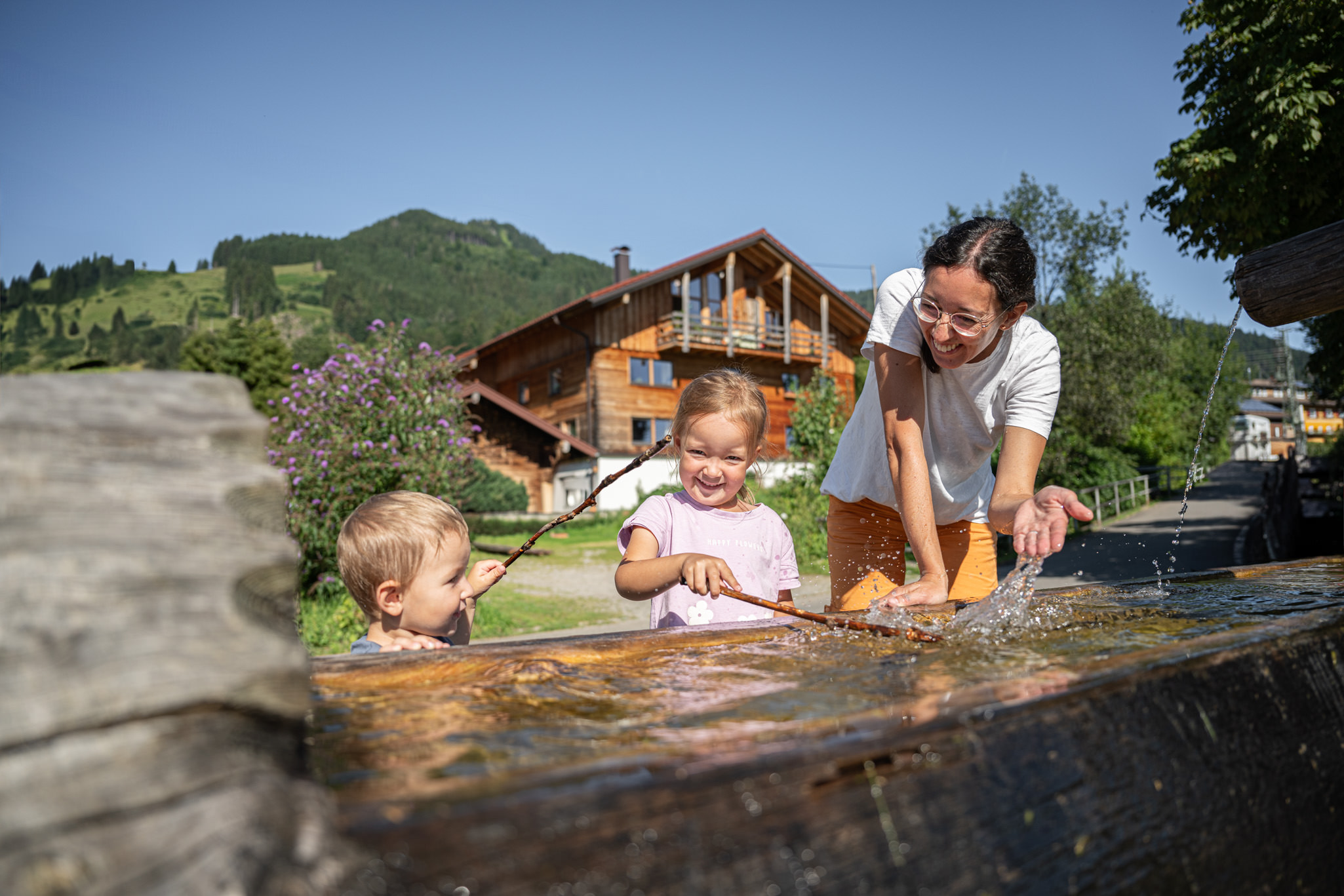 Mutter und zwei kleine Kinder erfrischen sich an einem Holzbrunnen, im Hintergrund ein Holzhaus, satte grüne Wiesen und blauer Himmel in Bolsterlang.