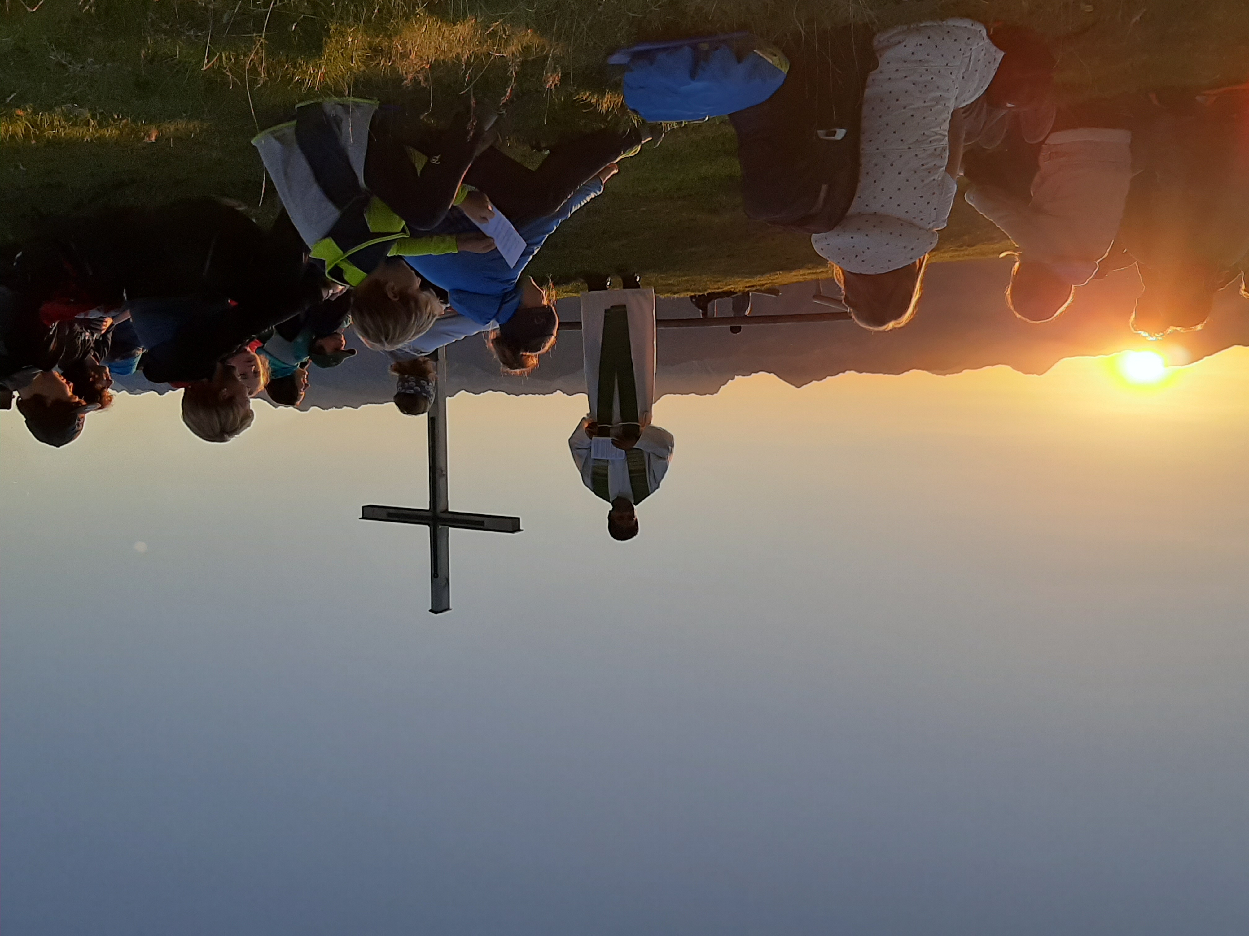 Bergmesse am Ofterschwanger Horn beim Sonnenaufgang. Ein Pfarrer vor Gipfelkreuz, umgeben von Gläubigen auf Bergwiese.