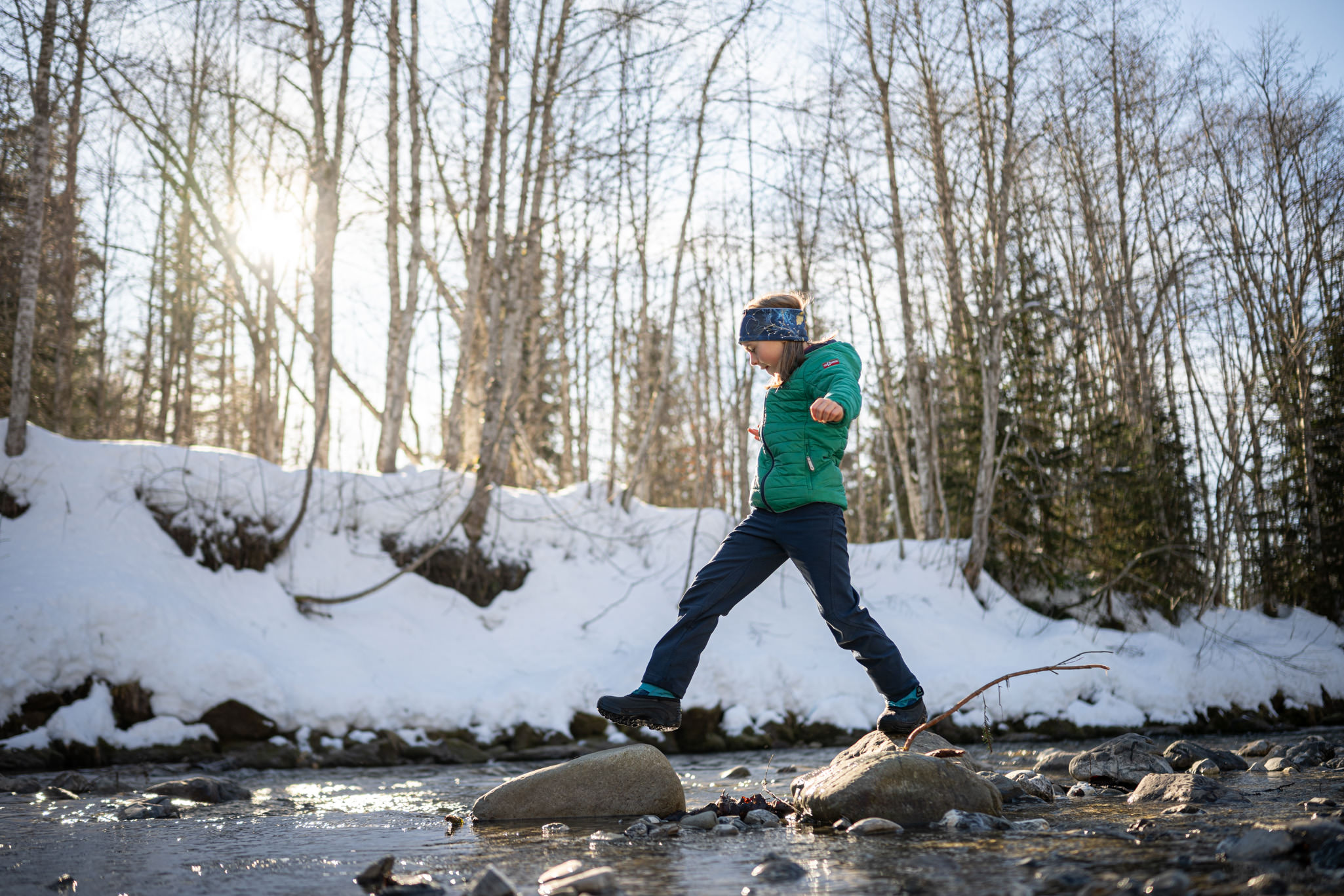Kind balanciert über Steine im Bach, im Hintergrund verschneiter Wald