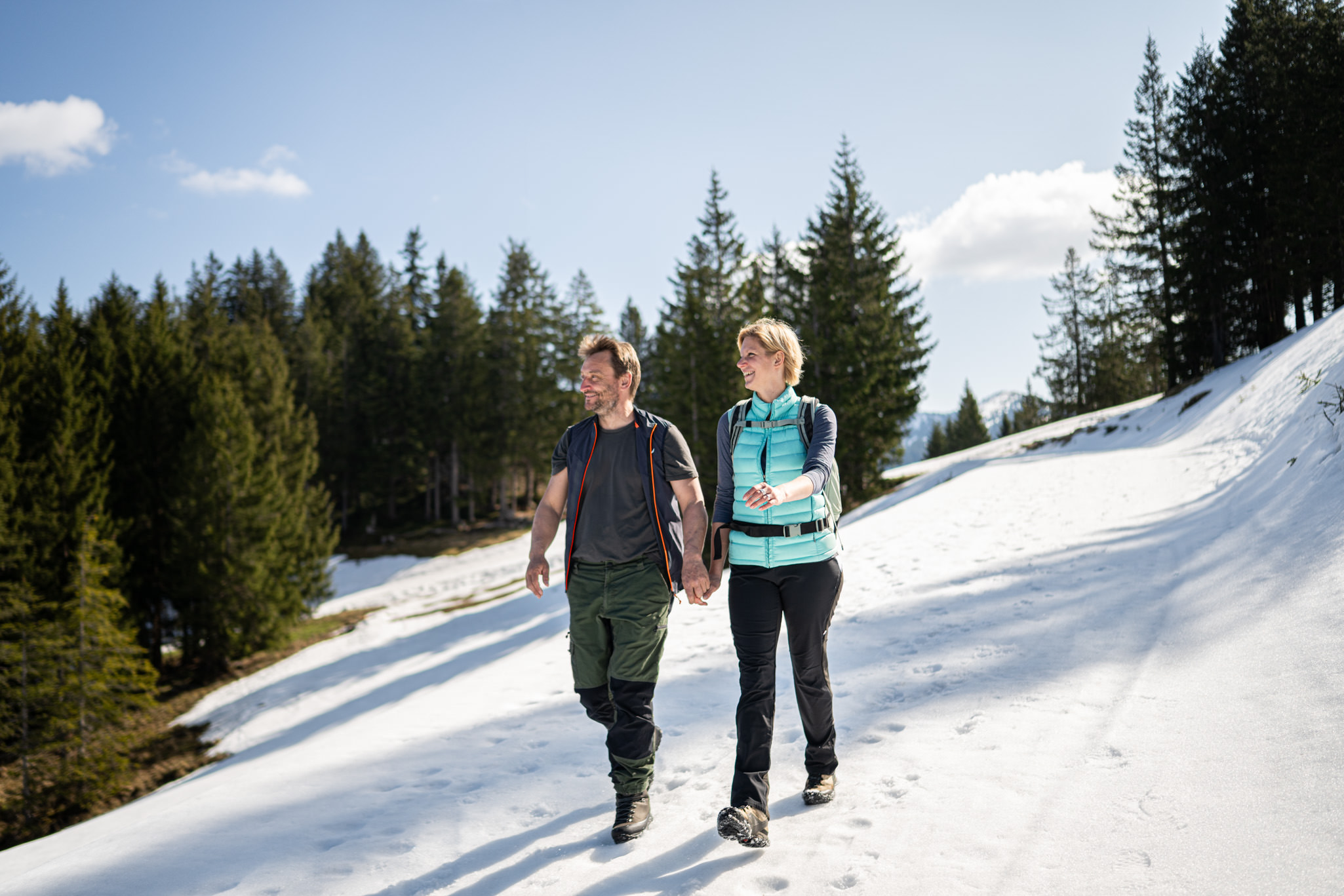Paar wandert auf verschneitem Weg, im Hintergrund grüner Tannenwald