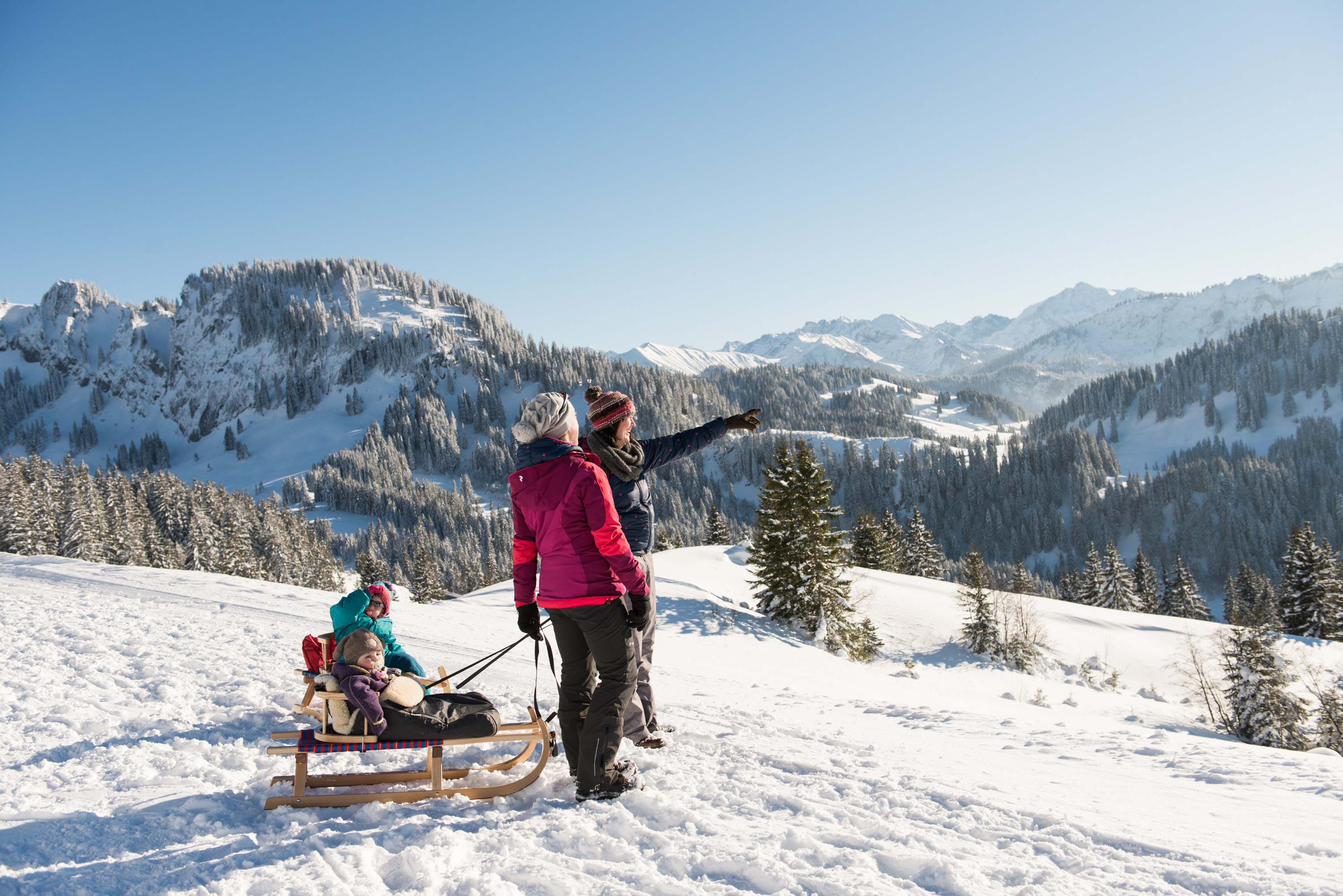 Zwei Frauen ziehen Kinder auf der dem Schlitten und bestaunen die verschneiten, sonnenbeschienenen Berge