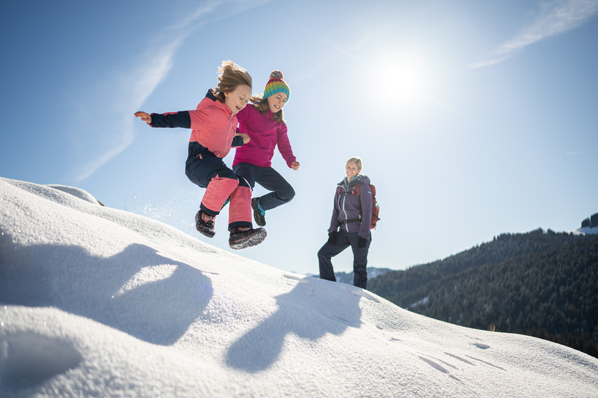 Zwei Kinder springen bei strahlendem Sonnenschein und blauem Himmel von einem Schneehügel.