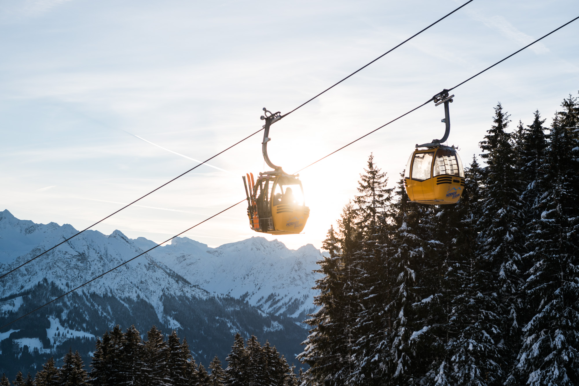 Die Hörnerbahn Bolsterlang in den Hörnerdörfern im Allgäu: Zwei Gondeln schweben vor verschneiten Allgäuer Alpen und dunklen Tannenwipfeln.
