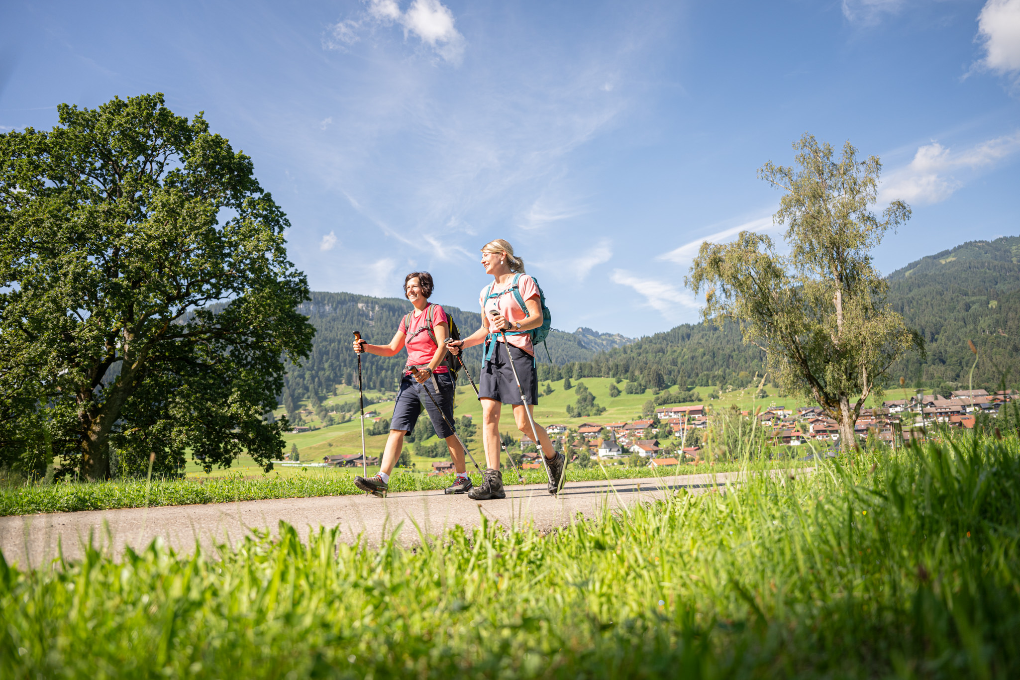 Zwei Wanderer auf sonnigen Weg durch grüne Wiesenlandschaft. Im Hintergrund der Ort Obermaiselstein in den Hörnerdörfern.