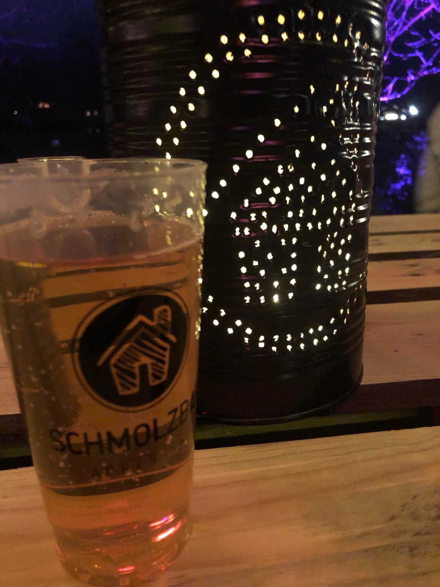 Ein Glas helles Getränk mit dem Aufdruck "SCHMOLZBAR" steht auf einem Holztisch. Dahinter eine dunkle Laterne mit ausgestanzten Mustern.