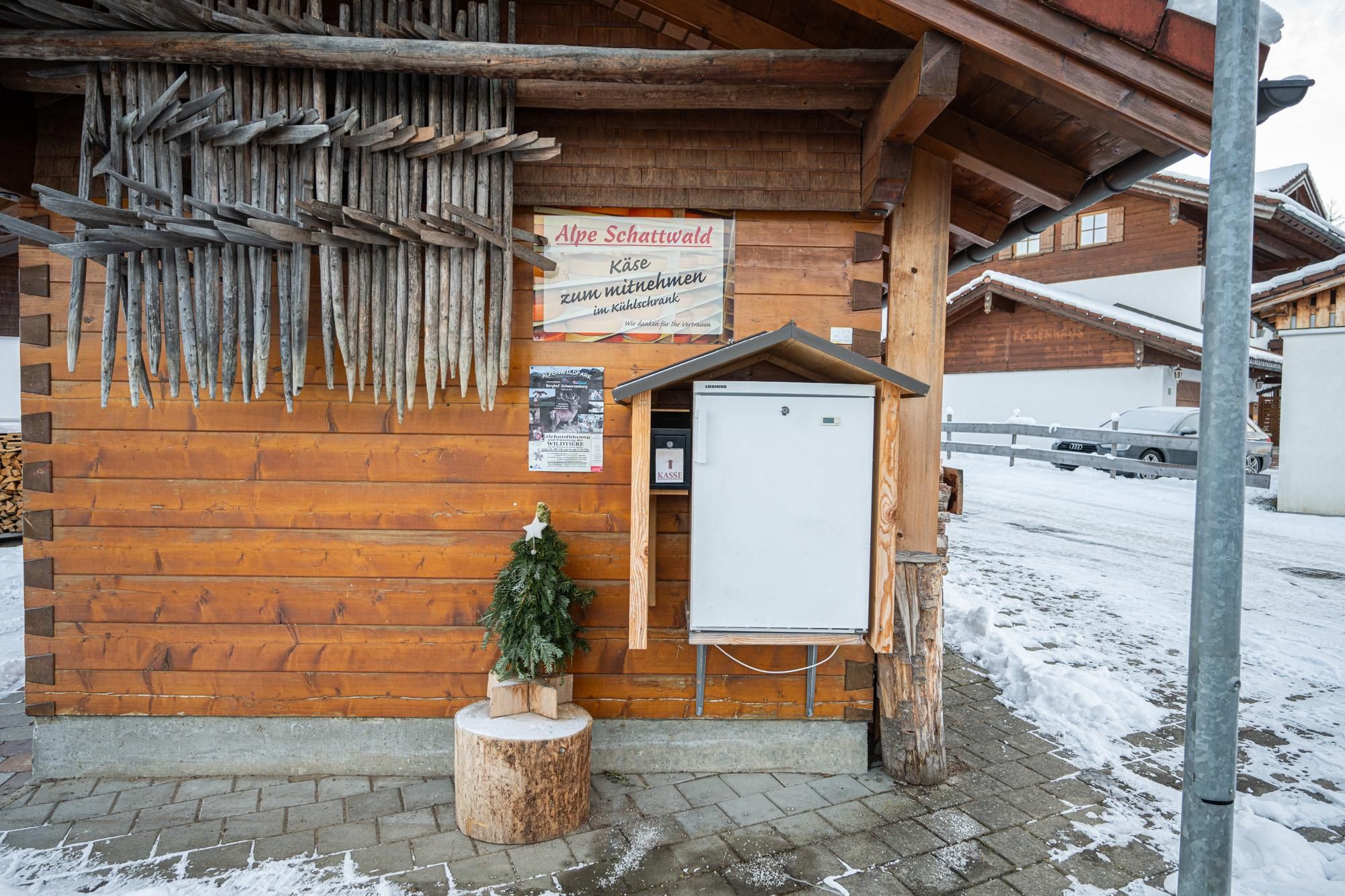 Hölzerne Hütte mit einem weißen Kühlschrank für Käse zur Selbstbedienung. Ein kleiner Weihnachtsbaum steht davor. Schnee liegt auf dem Boden.