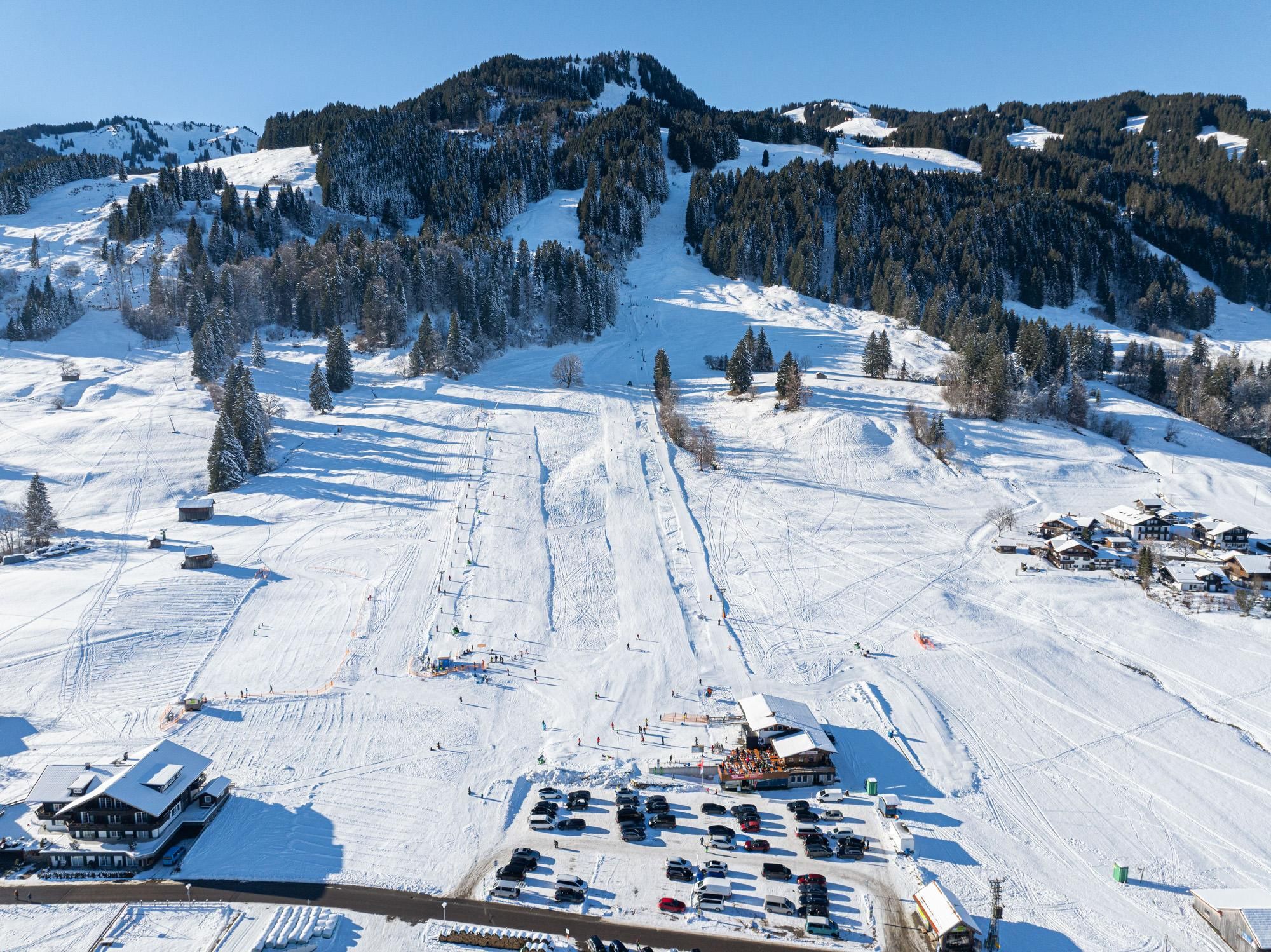 Verschneite Pisten, Skilifte, Gebäude, Parkplatz mit Autos, bewaldete Hänge, blauer Himmel.