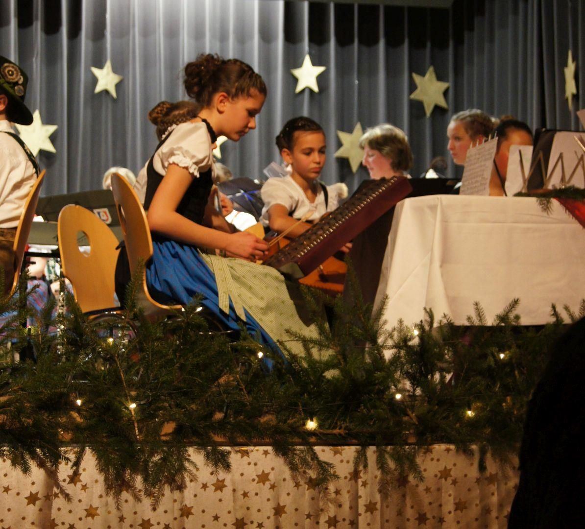 Junge Frauen in Tracht spielen Hackbrett auf einer Bühne. Festliche Dekoration mit Lichtern und Sternen. Vorhang im Hintergrund.
