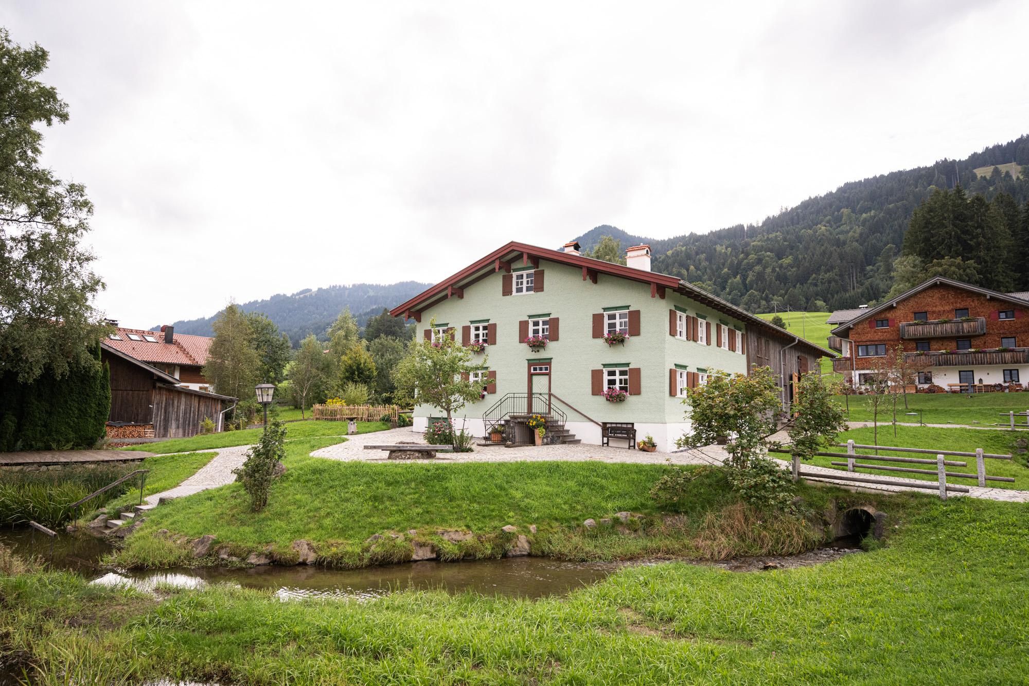 Helles Haus mit braunen Fensterläden und rotem Dach, umgeben von grüner Wiese und einem kleinen Fluss. Ein Gehweg führt zum Haus. Bewaldete Hügel im Hintergrund.