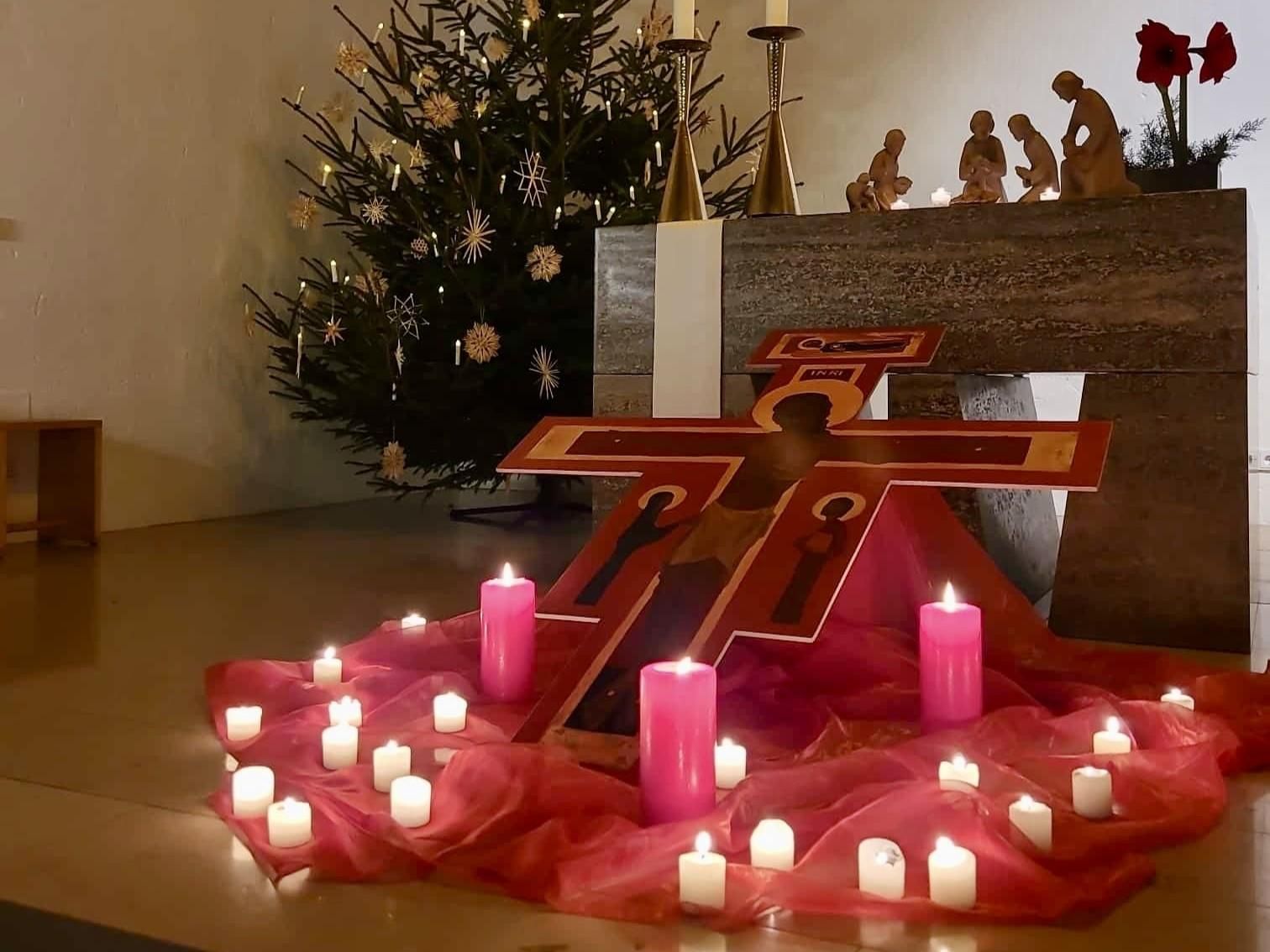Ein dekorierter Altar mit einem Kreuz und vielen Kerzen. Im Hintergrund steht ein Weihnachtsbaum und eine Krippe steht auf dem Tisch.