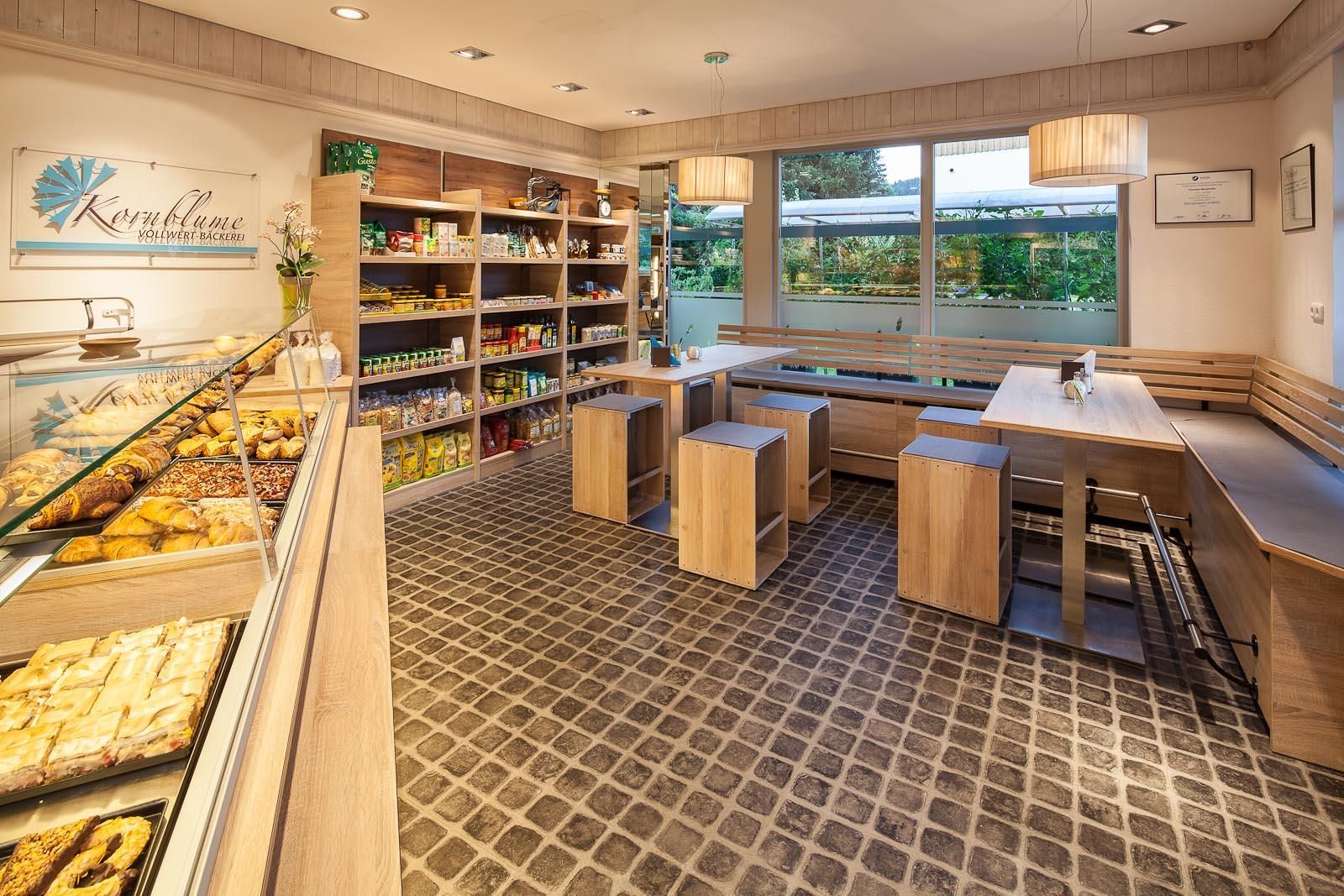 Helle Bäckerei mit Verkaufstheke links. Regale mit Produkten an der Wand. Sitzbereich mit Tischen und Bänken rechts.