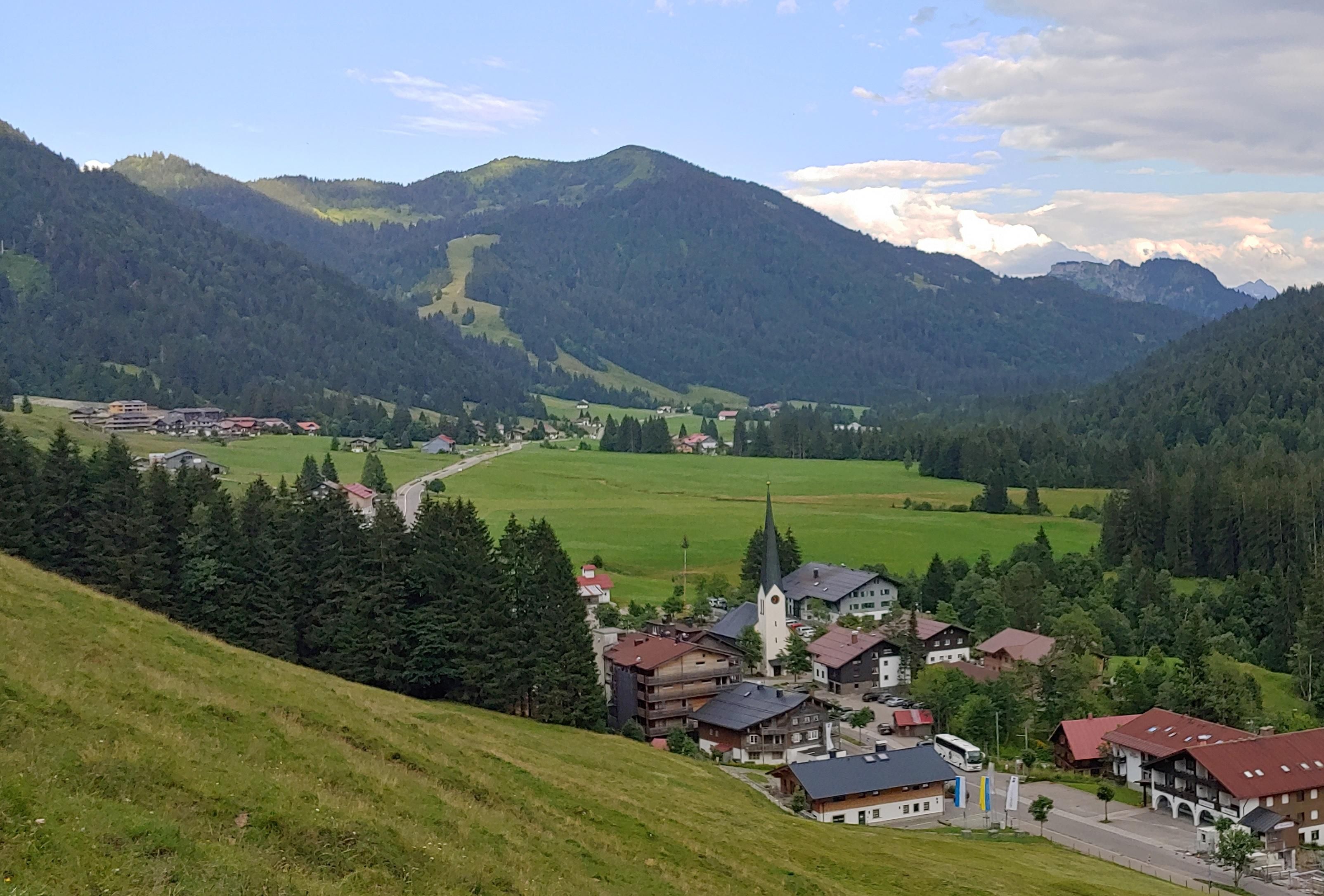 Panorama von Balderschwang heute. Ein Dorf mit dunklen Dächern liegt in einem grünen Tal, umgeben von bewaldeten Bergen.