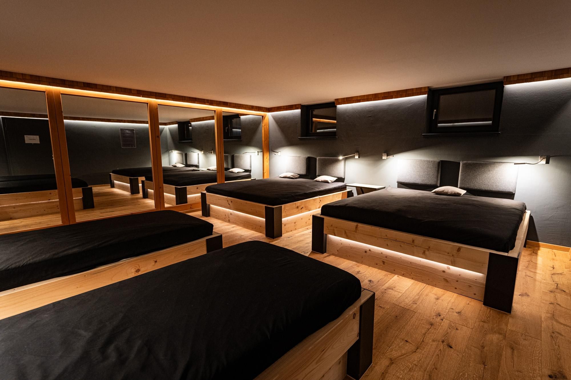 Heller Ruheraum.  Mehrere Holzbettgestelle mit schwarzen Auflagen. Indirekte Beleuchtung unter den Betten und an den Wänden.