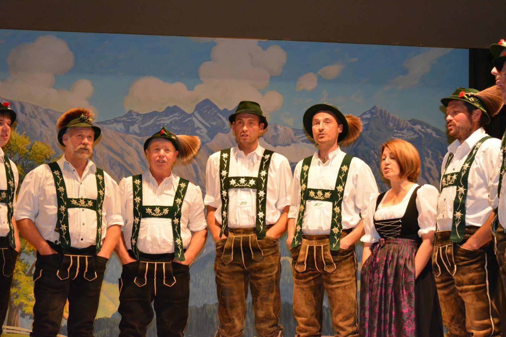 Gruppe an Menschen stehen auf einer Bühne und singen. Alle haben die traditionelle Tracht an.