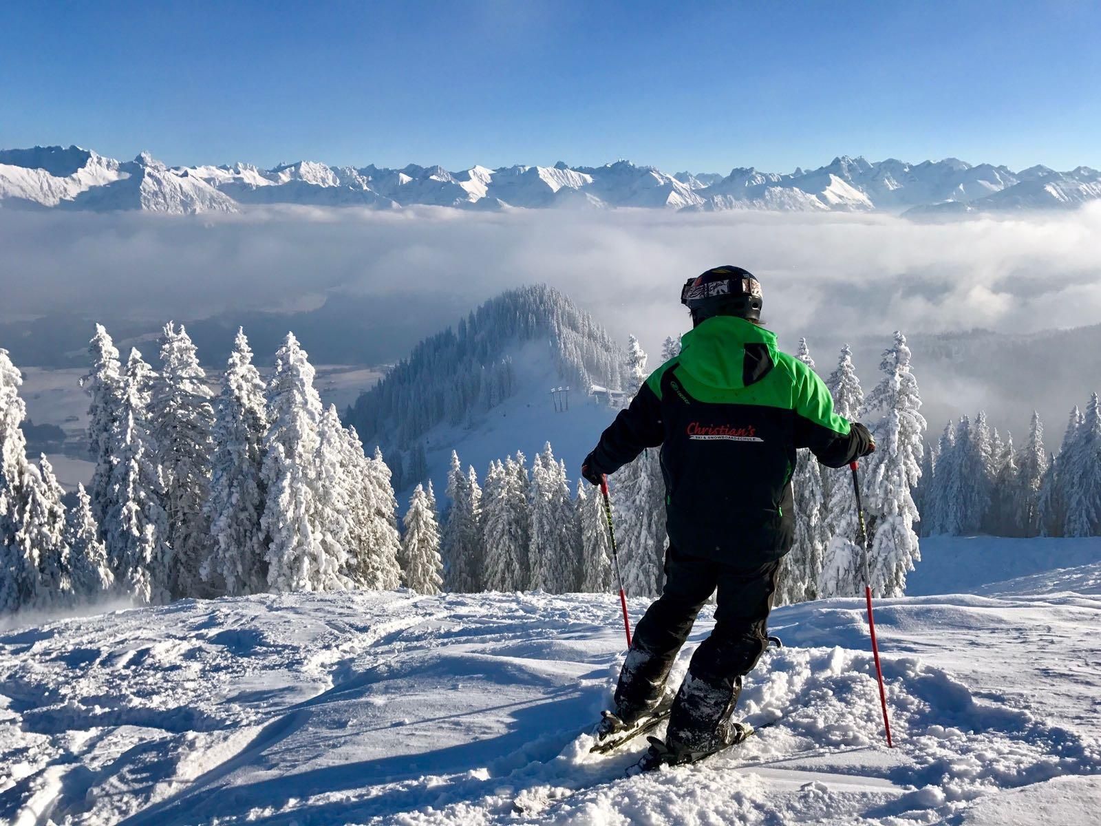 Ein Skilehrer steht auf einem schneebedeckten Berg und schaut auf die beeindruckende Berglandschaft. Der Himmel ist klar und es gibt niedrige Wolken über den Bergen.