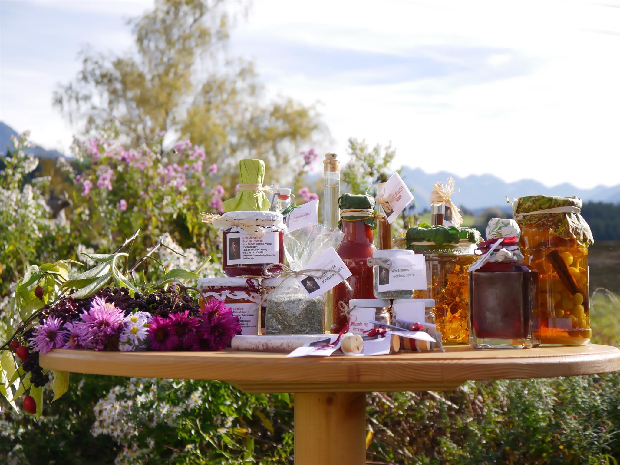 Runder Holztisch im Freien mit verschiedenen Kräuterprodukten in Gläsern und Flaschen. Blumen und Pflanzen umgeben den Tisch. Berge im Hintergrund.