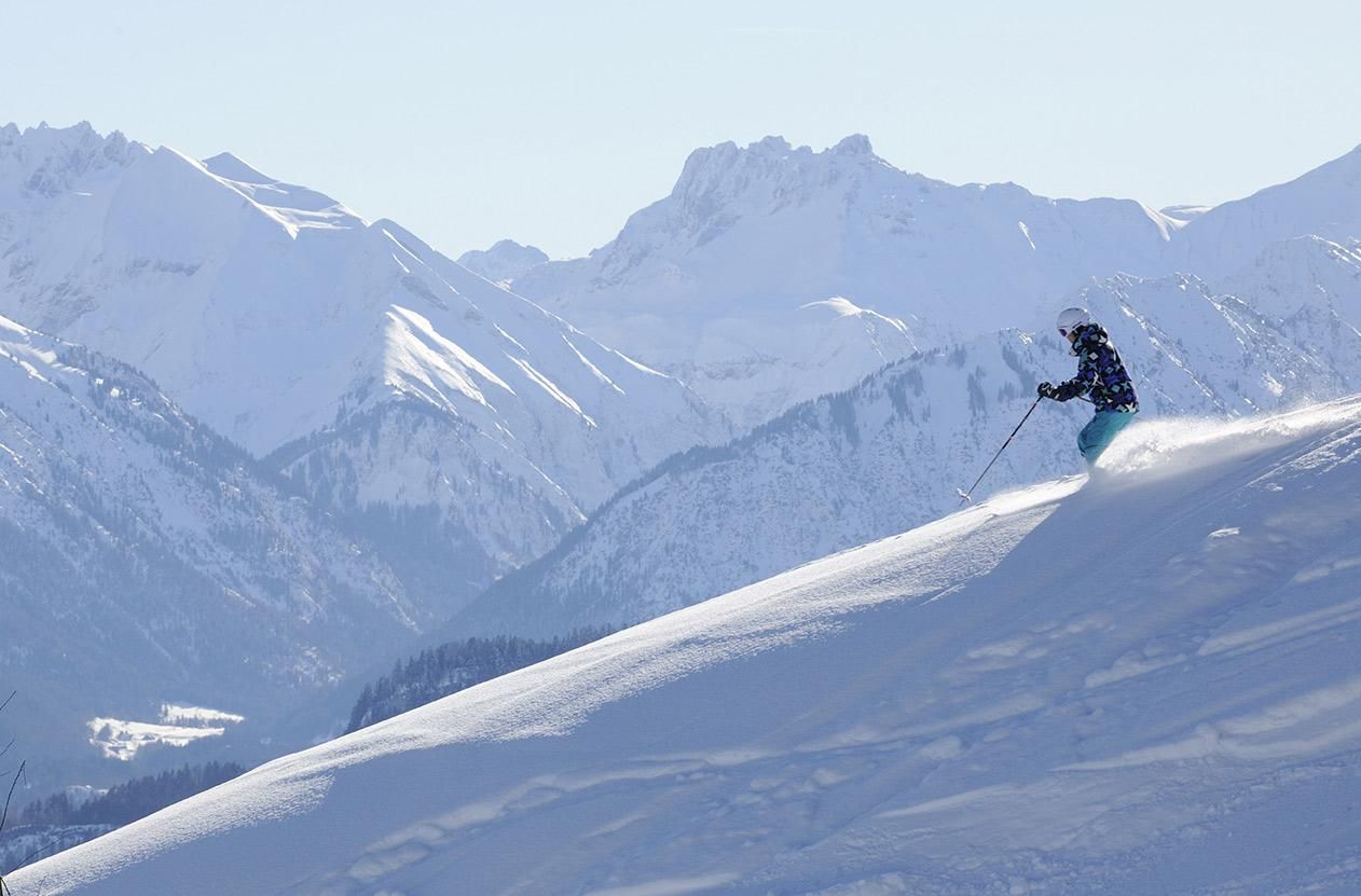 Skifahrerin in gemusterter Jacke auf einer steilen, verschneiten Piste. Majestätische Bergkulisse im Hintergrund.