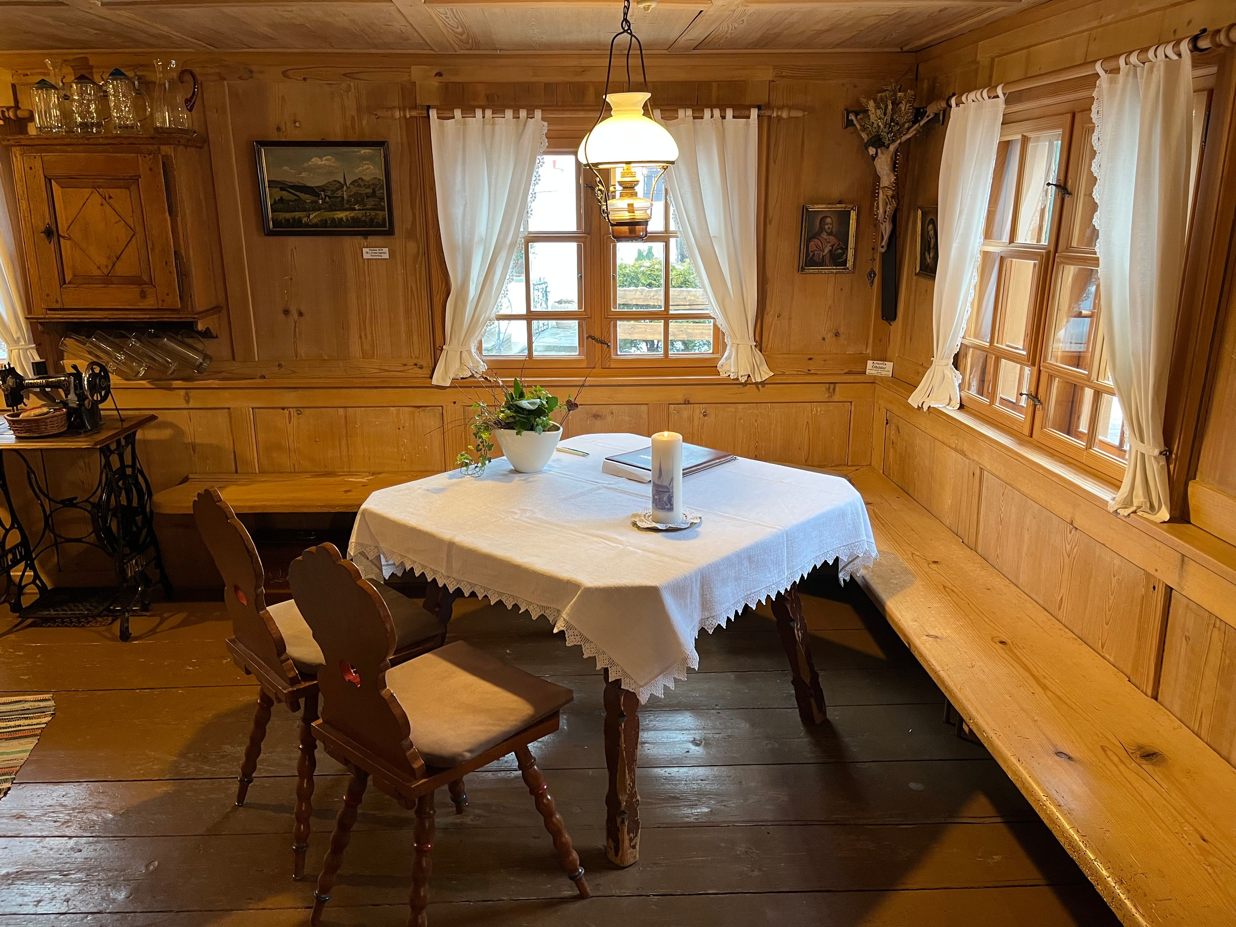 Bauernstube im alpenländischen Stil, Holztisch mit Tischdecke, Bänke und Stühle, Fenster mit Vorhängen.