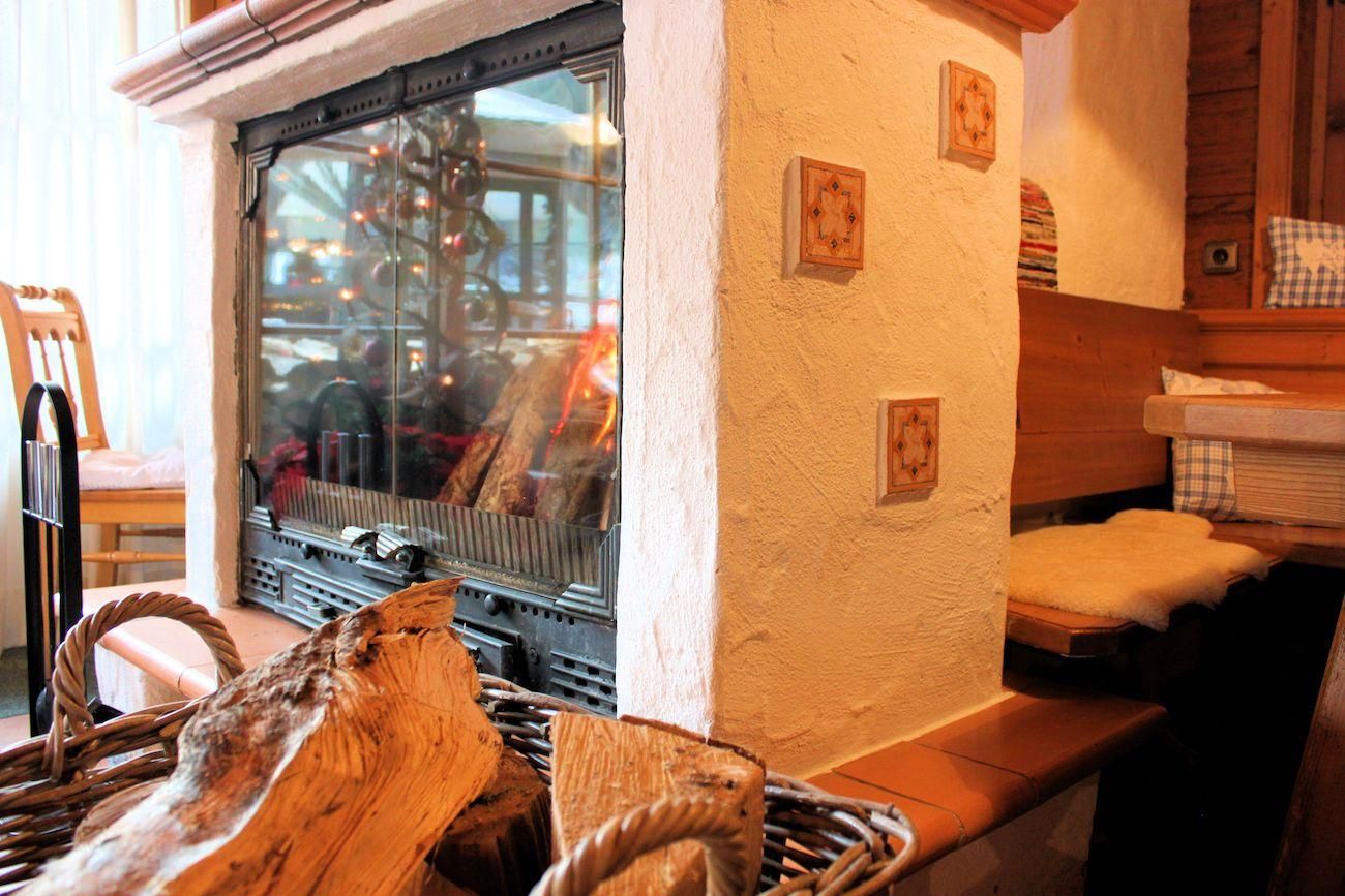 Gemütlicher Kaminbereich. Weißer Kamin mit Glasscheibe. Korb mit Holzscheiten davor. Holzvertäfelte Wände und eine Eckbank mit Kissen im Hintergrund.