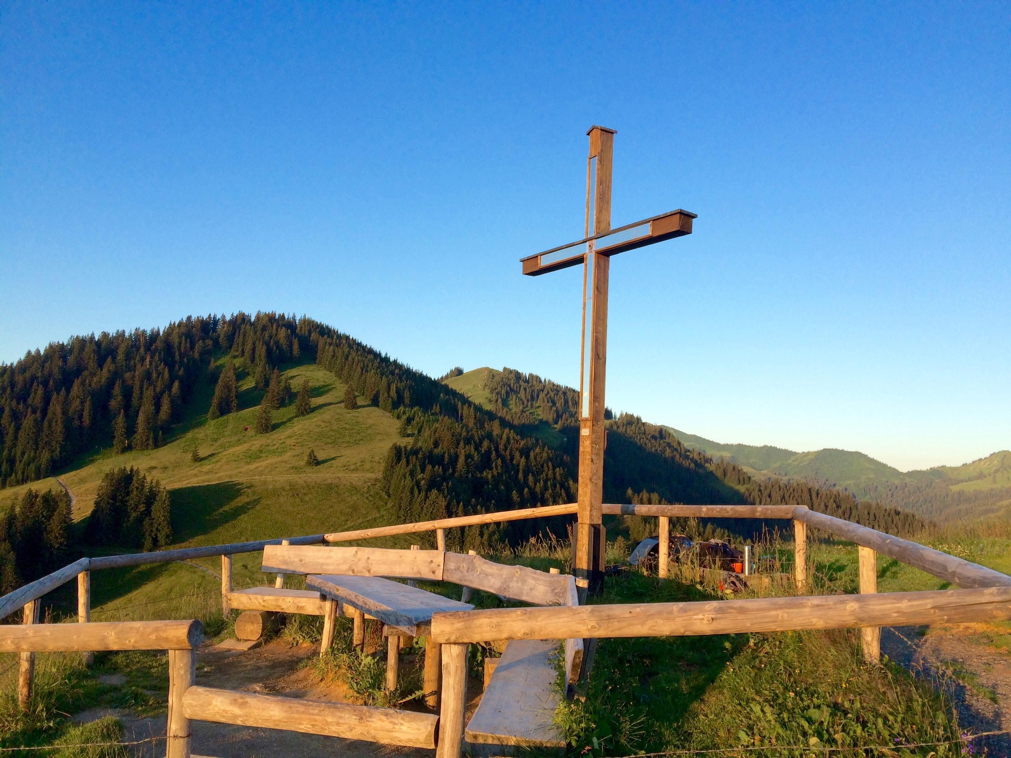 Ein Gipfelkreuz aus Holz auf einem Bergkamm, umgeben von grünen Hügeln und Wäldern unter blauem Himmel.