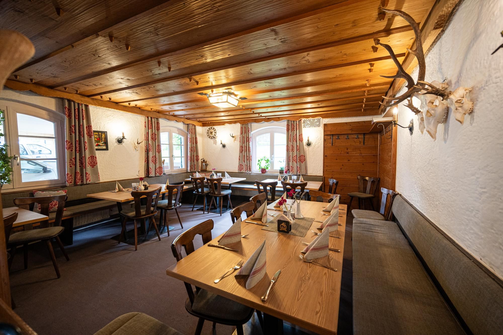 Holzvertäfelte Decke und teilweise holzverkleidete Wände. Helle Holztische mit Gedeck und Stühlen sowie Eckbänken. Dekoriert mit Hirschgeweih. Fenster mit rot gemusterten Vorhängen.