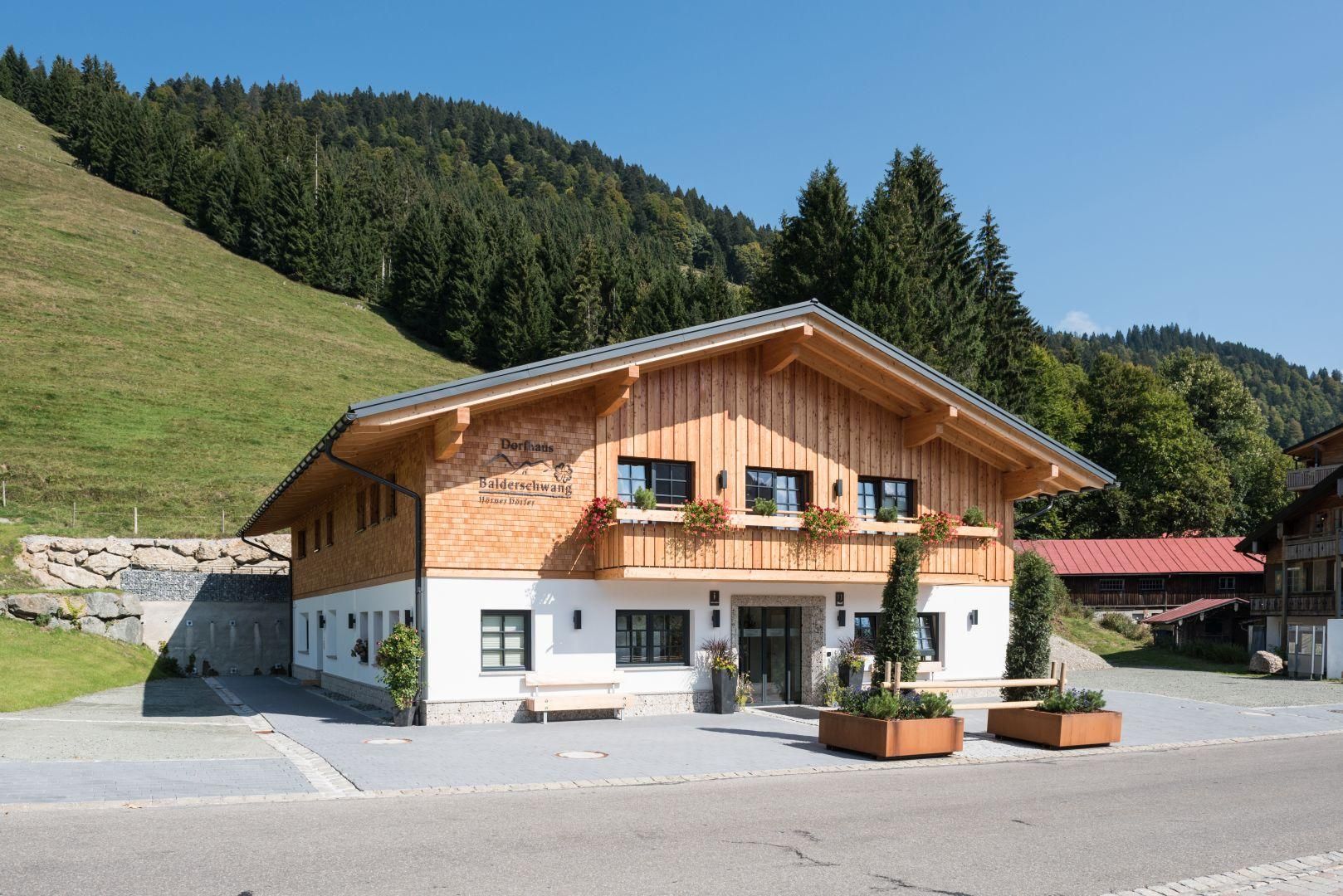 Dorfhaus mit Holzfassade und weißem Erdgeschoss. Balkonkästen. Im Hintergrund grüne Berge.