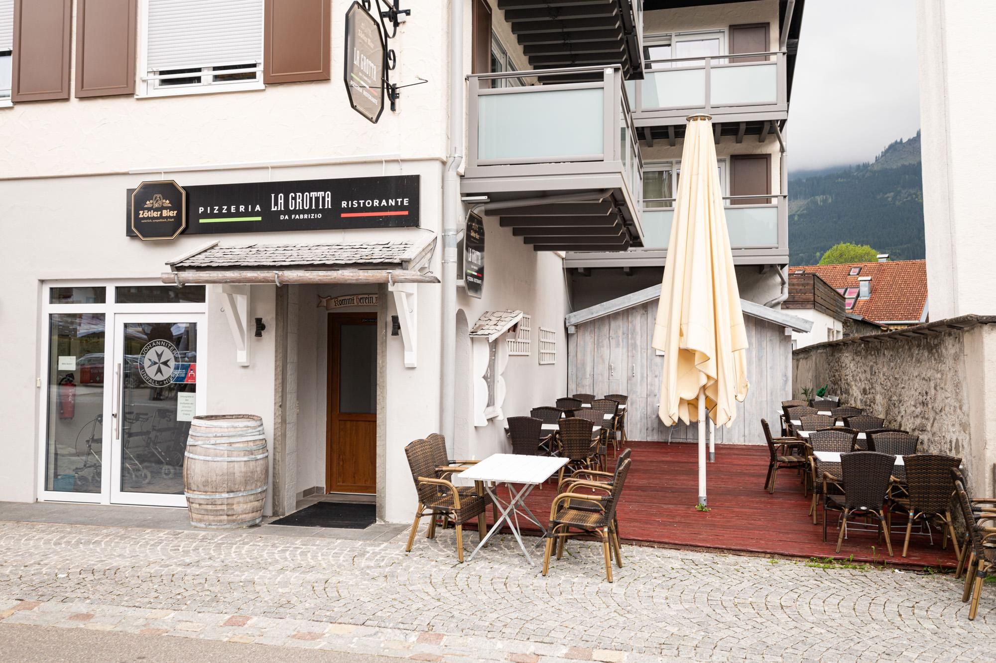 Eingang zur Pizzeria "La Grotta", rechts der Außenbereich mit Tischen und großem, hellgelben Schirm.