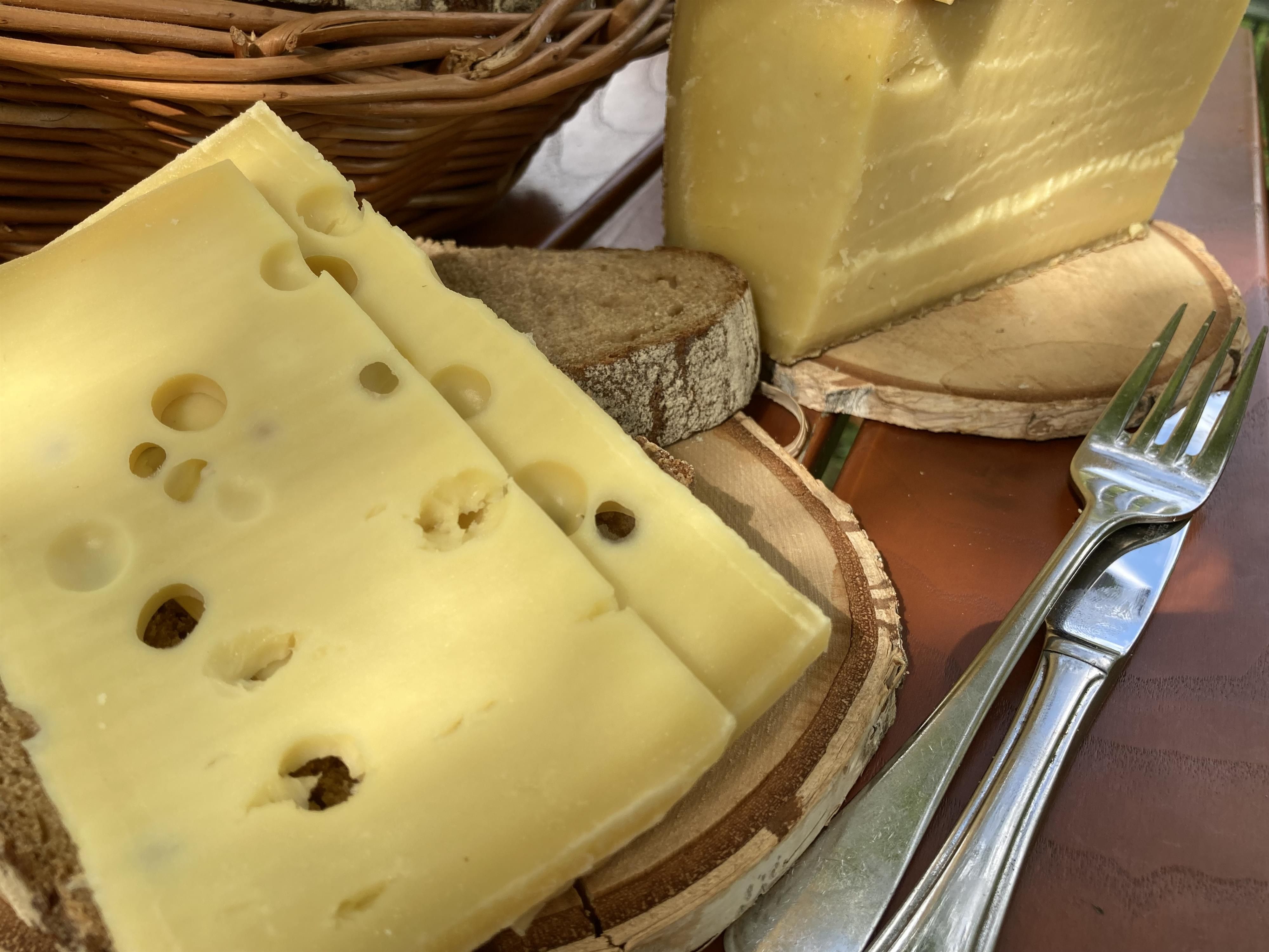 Drei Scheiben löchriger Käse auf Brot, dazu Messer und Gabel. Im Hintergrund ein Stück Käse und ein Korb.