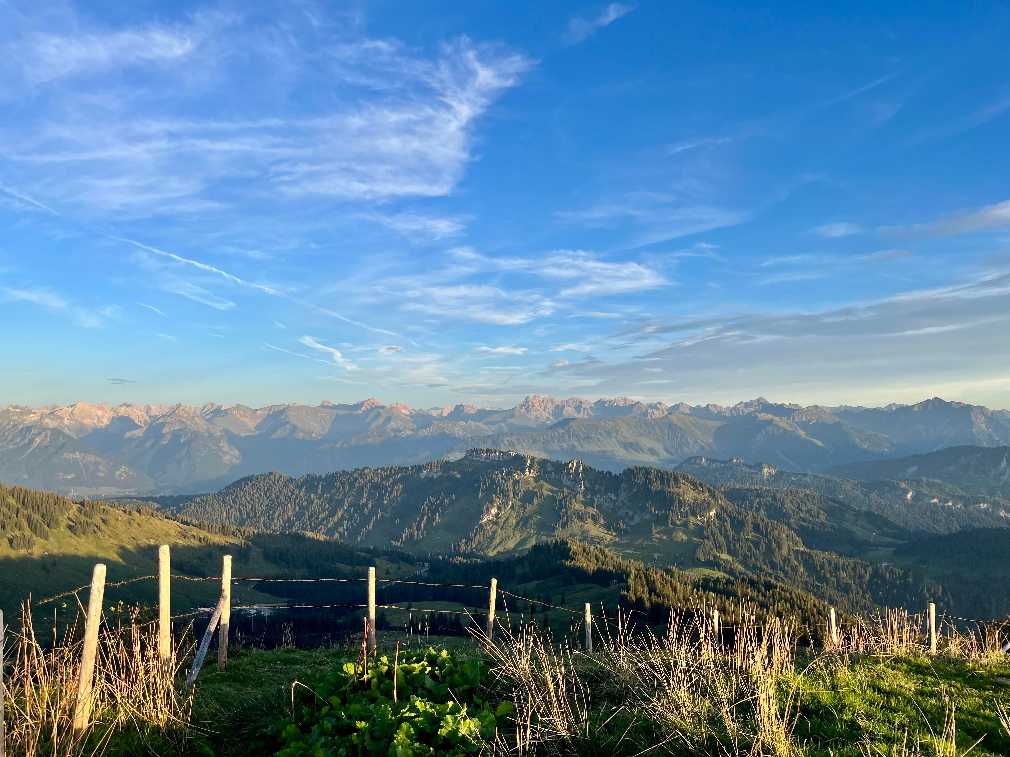 Eine Berglandschaft mit malerischen Bergen und einem klaren blauen Himmel. Im Vordergrund sind vereinzelte Zäune und grünes Gras zu sehen.