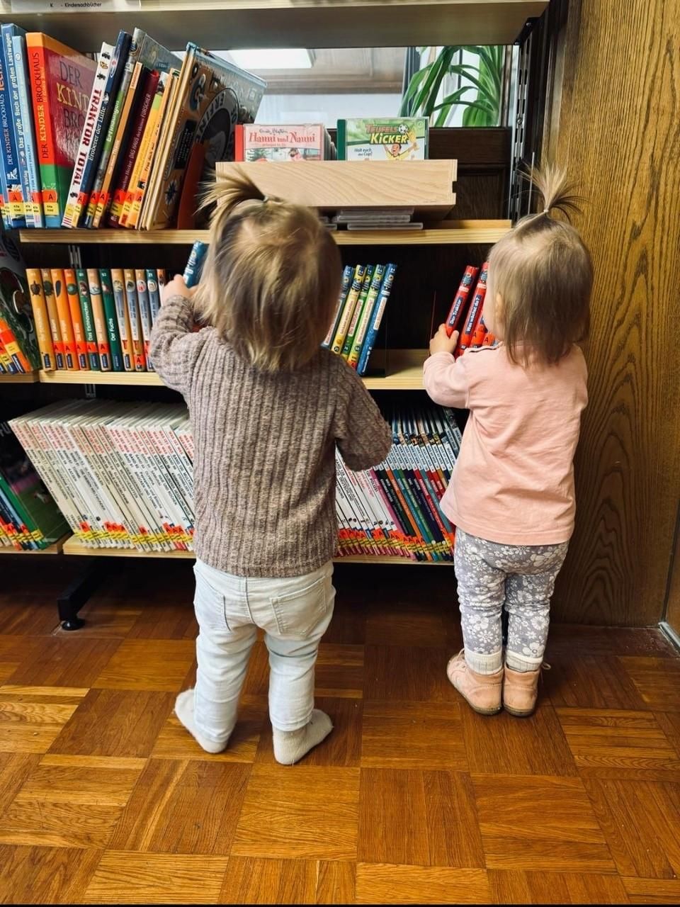 Zwei kleine Kinder stehen vor einem Bücherregal und schauen Bücher an.