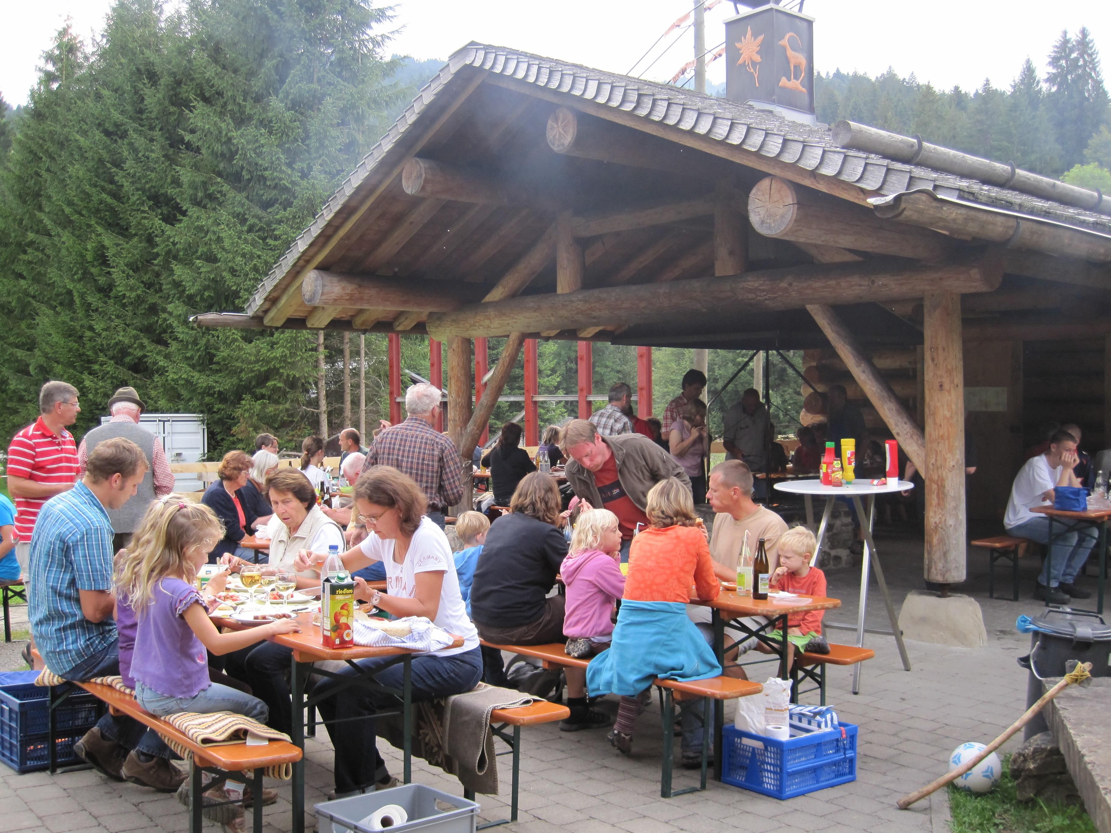 Ein großes Holzdach überdacht eine Terrasse mit Holztischen und Bänken, an denen viele Menschen sitzen und essen. Rauch steigt von einem Grill auf.