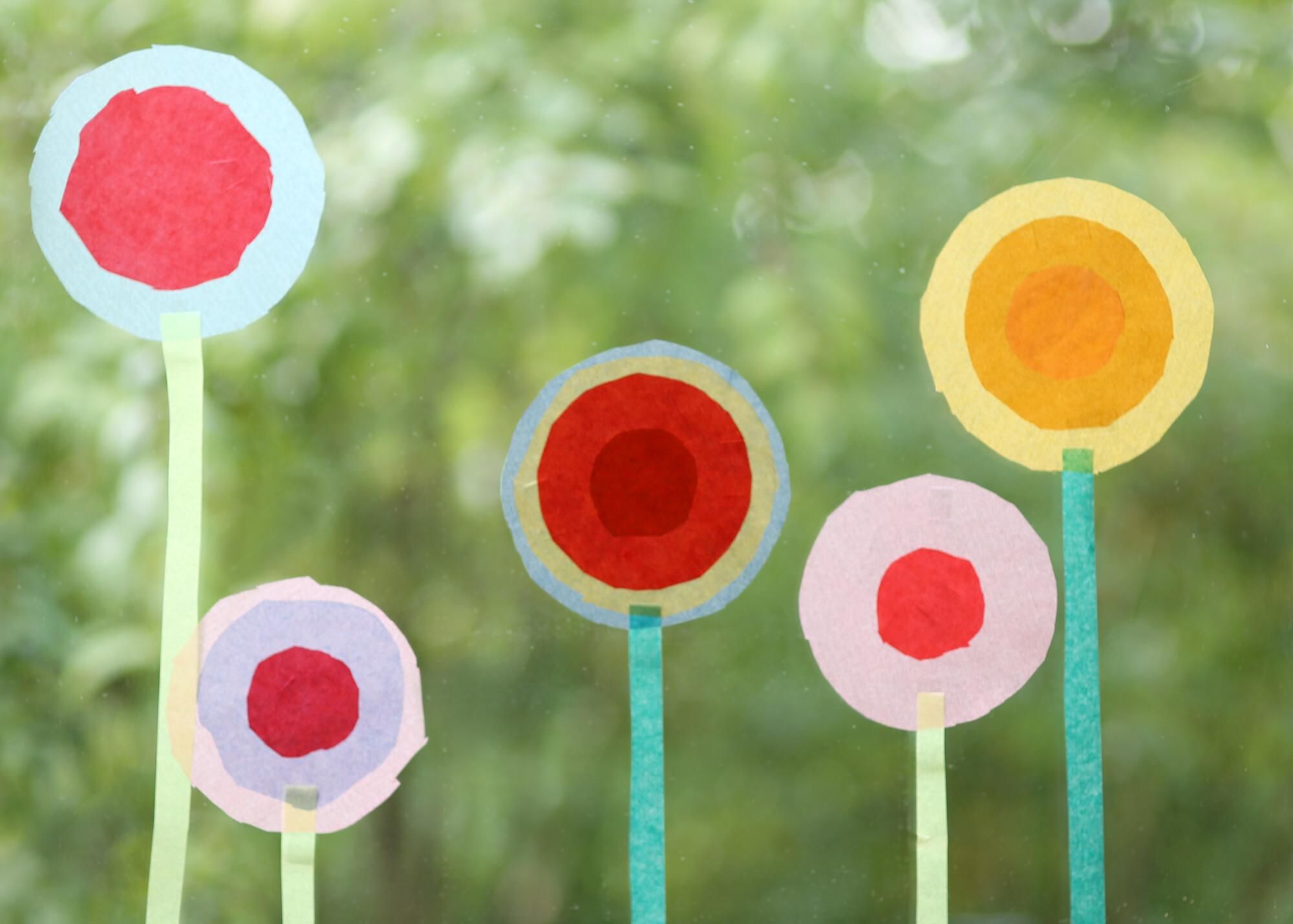 Bunte Blumen aus Papier stehen vor einem verschwommenen grünen Hintergrund. Sie haben verschiedene Farben und Formen, die eine fröhliche Stimmung vermitteln.